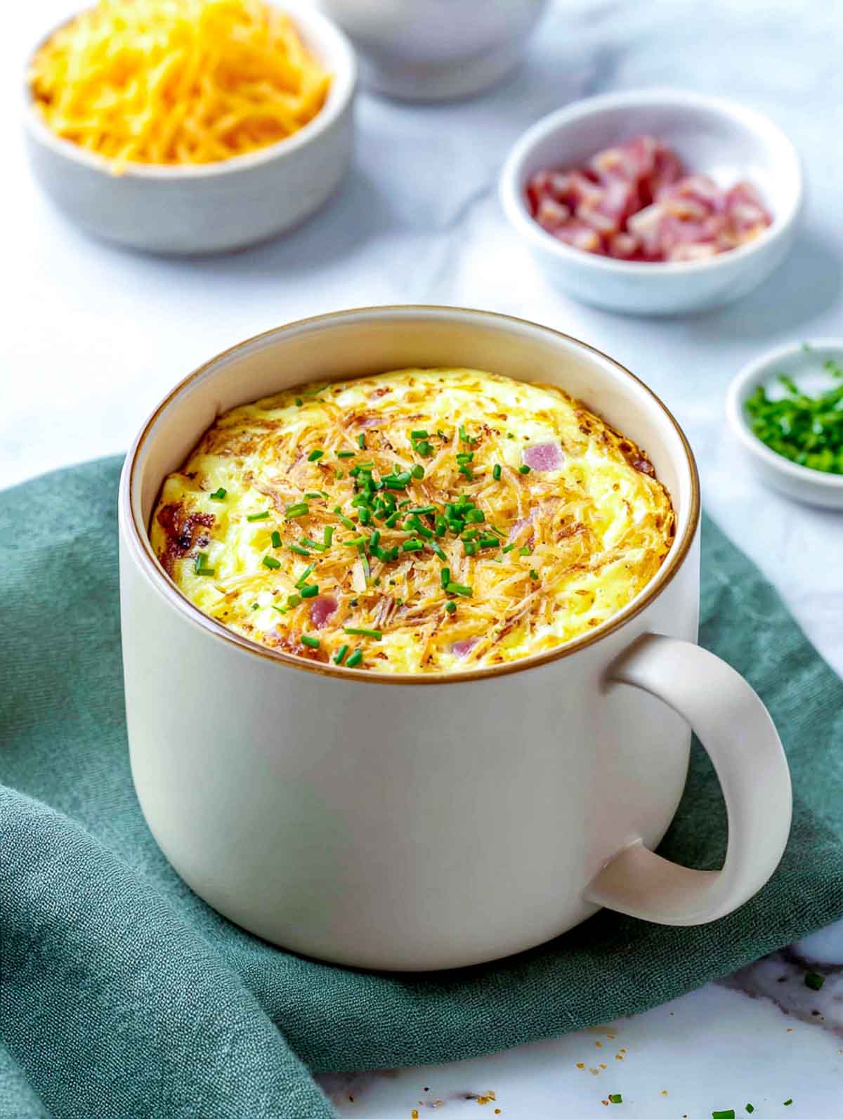 A cream ceramic mug filled with a fluffy egg bake mixed with diced ham and Swiss cheese, topped with a thin golden layer of shredded hash browns and chopped chives, set on a white marble countertop with a green napkin nearby.