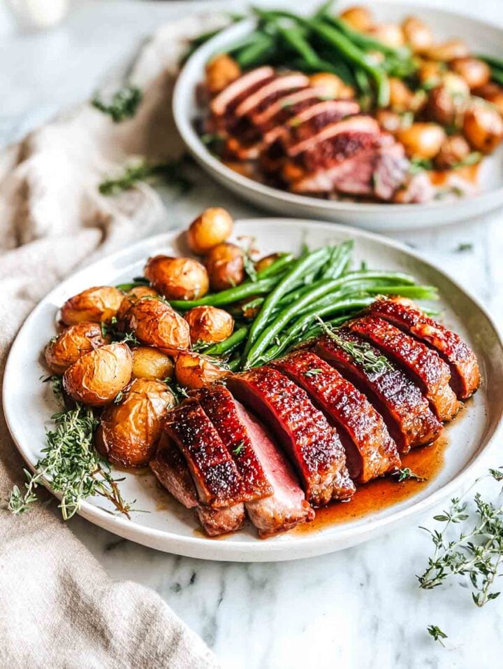 Two plates of sliced duck breast with roasted potatoes and green beans.