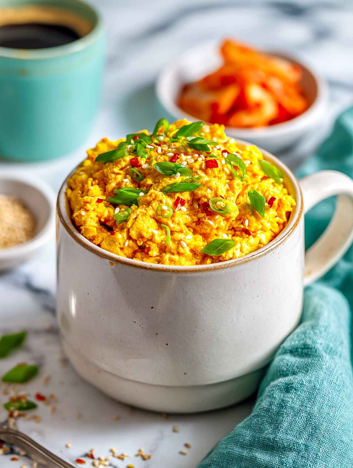 A light ceramic mug filled with golden tofu scramble mixed with kimchi and topped with sliced green onions and sesame seeds, set on a white marble countertop with a teal napkin and small bowls of sesame seeds and kimchi nearby.