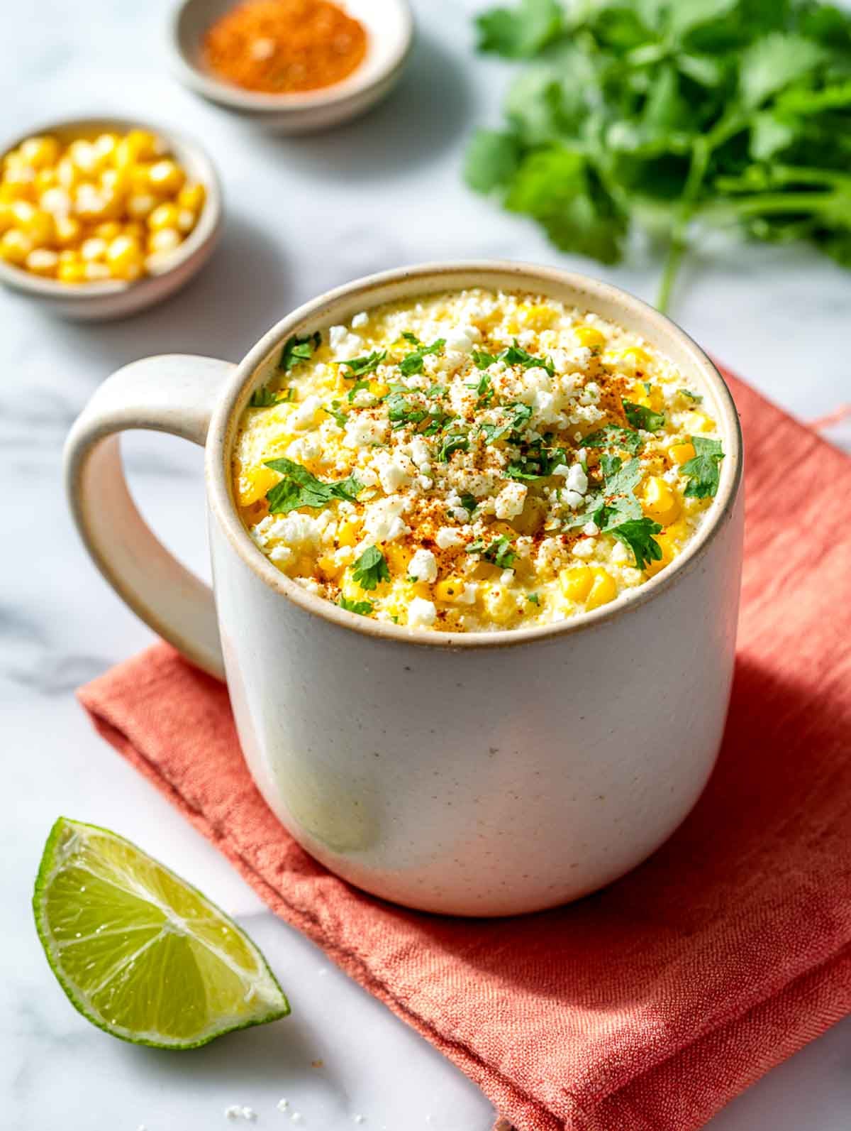 A bright angled side view of a white ceramic mug filled with fluffy eggs mixed with sweet corn, topped with crumbled cotija cheese, chopped cilantro, and a sprinkle of chili powder, set on a white marble countertop with a coral linen napkin and a lime wedge nearby.