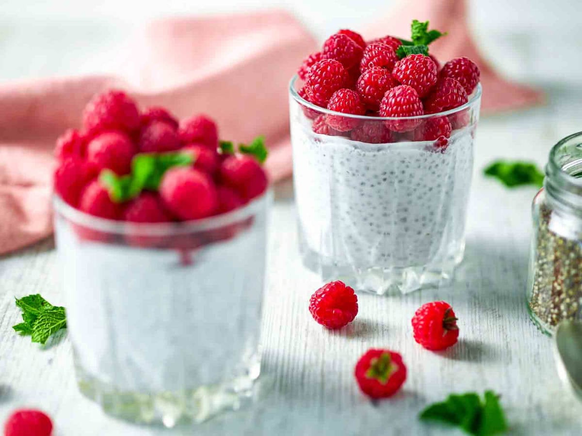 Two glasses of creamy chia pudding topped with fresh raspberries and mint on a light tabletop