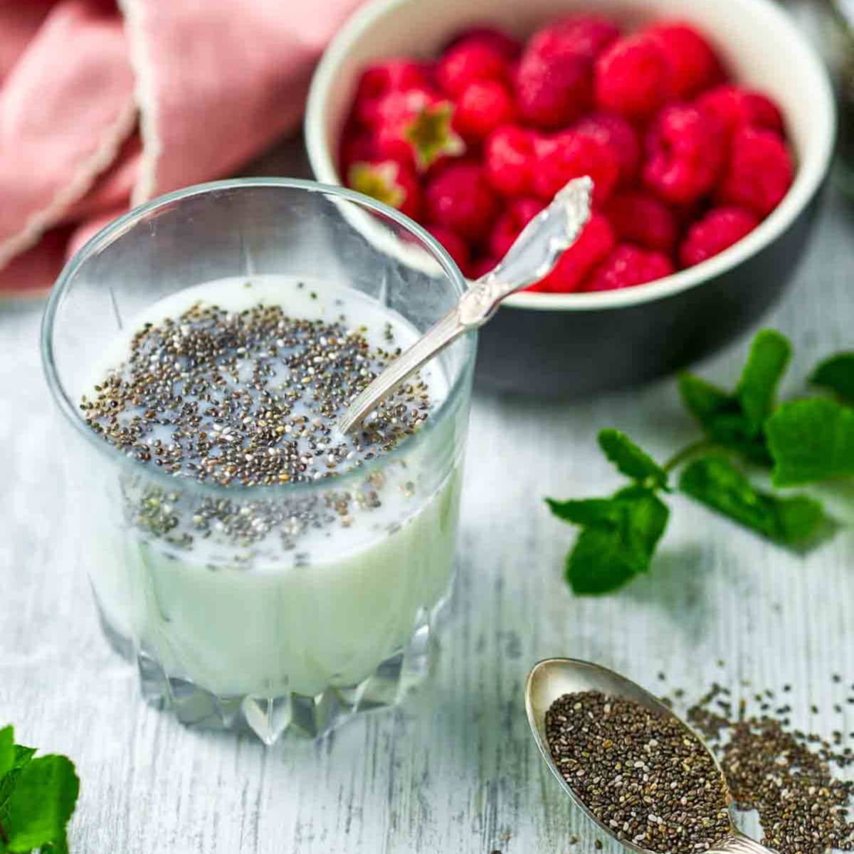 Glass of milk with chia seeds soaking, spoon inside, with fresh raspberries and mint nearby