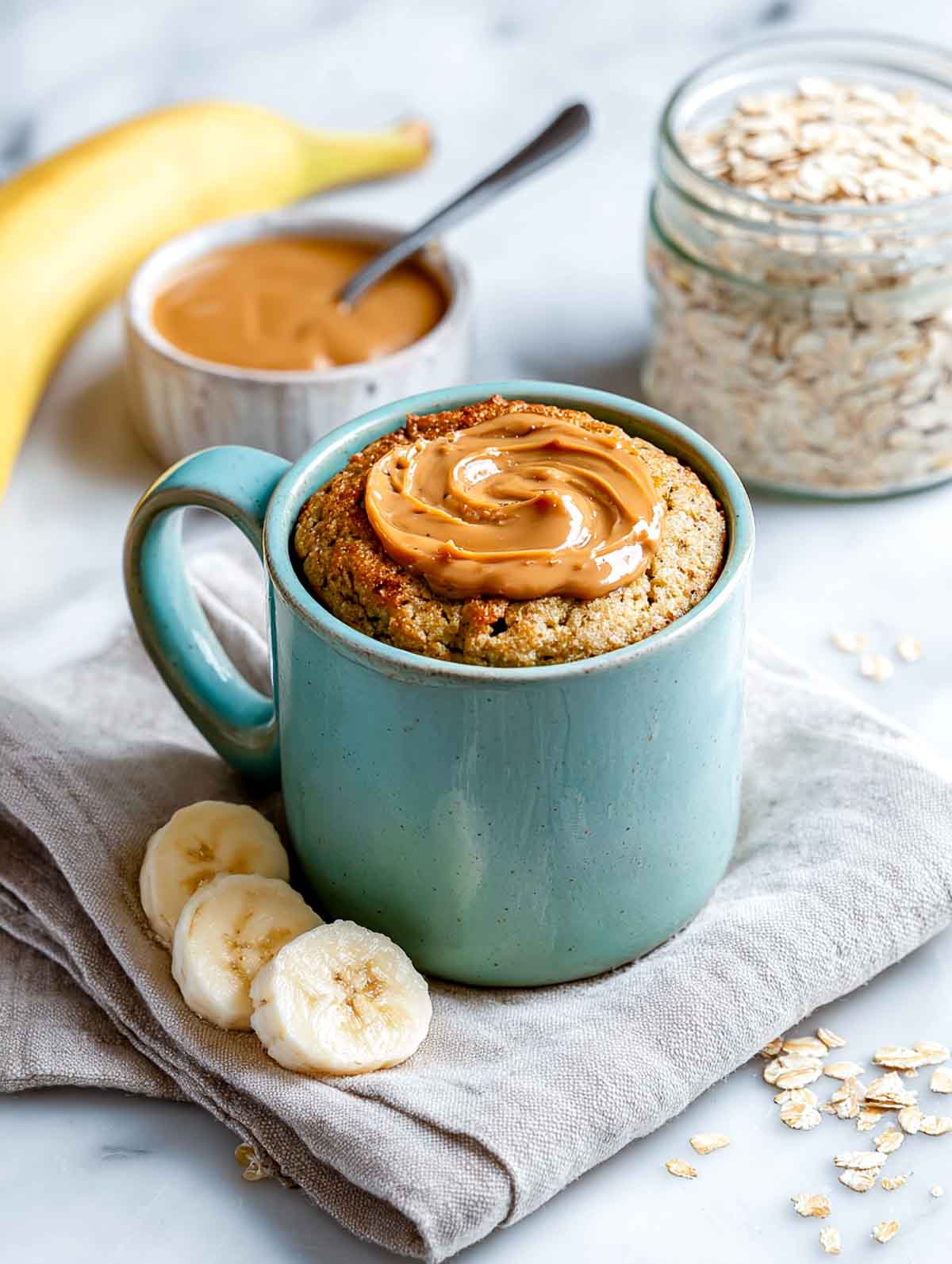 A bright angled view of a pale sage green mug filled with a peanut butter banana oat protein mug cake, topped with a thick peanut butter swirl, set on a white marble countertop with banana slices, a jar of oats, and a small bowl of peanut butter nearby.