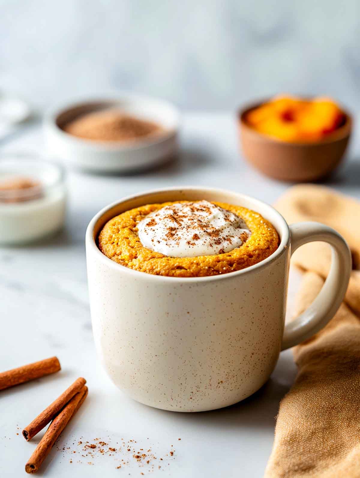 A cream ceramic mug filled with a warmly spiced pumpkin protein mug cake topped with a small dollop of yogurt and a light sprinkle of cinnamon, set on a white marble countertop with cinnamon sticks and small bowls of pumpkin and spices nearby.