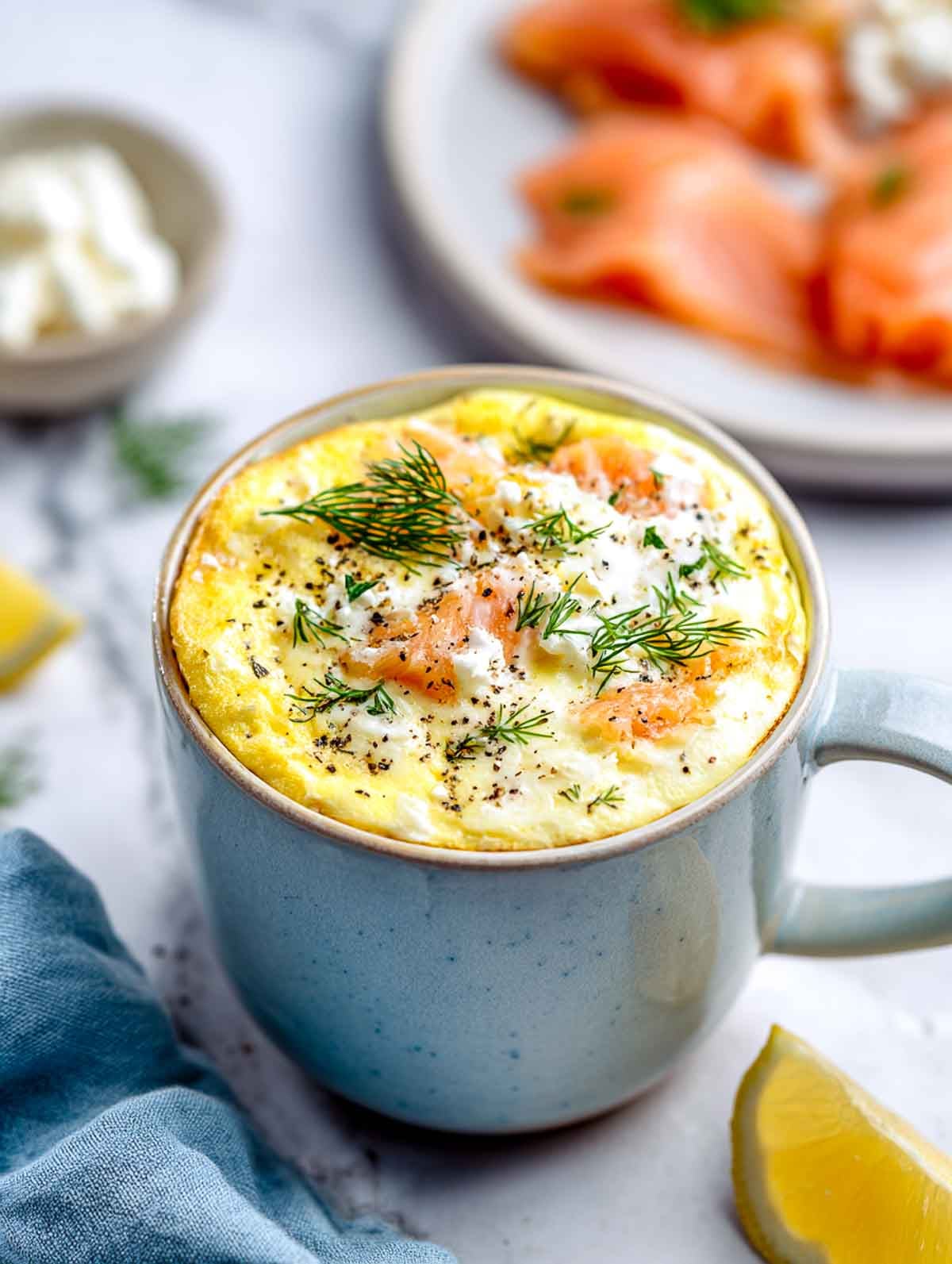 A light ceramic mug filled with a fluffy egg bake studded with smoked salmon and cottage cheese, topped with fresh dill and black pepper, set on a white marble countertop with lemon wedges and a blue napkin nearby.