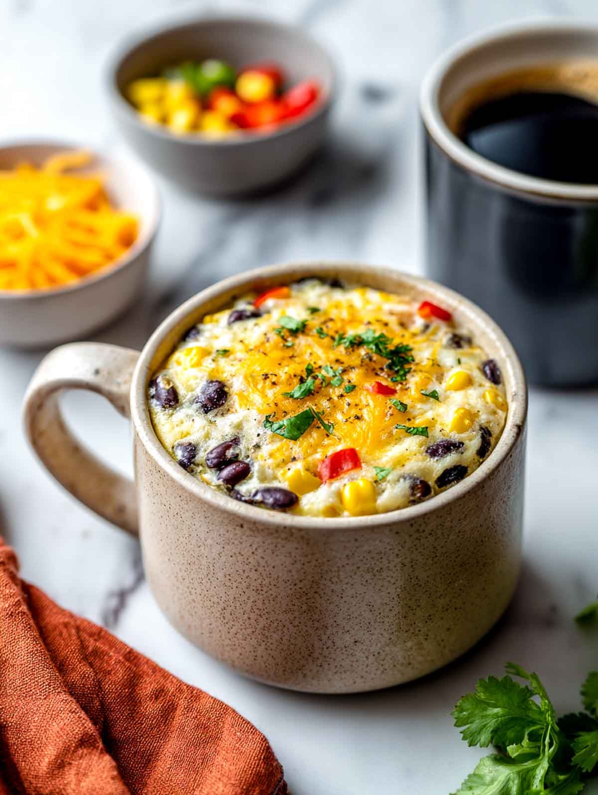 A sand-colored ceramic mug filled with a fluffy egg white bake studded with black beans, corn, and red peppers, topped with melted cheddar and chopped cilantro, set on a white marble countertop with a burnt orange napkin and small bowls of ingredients nearby.