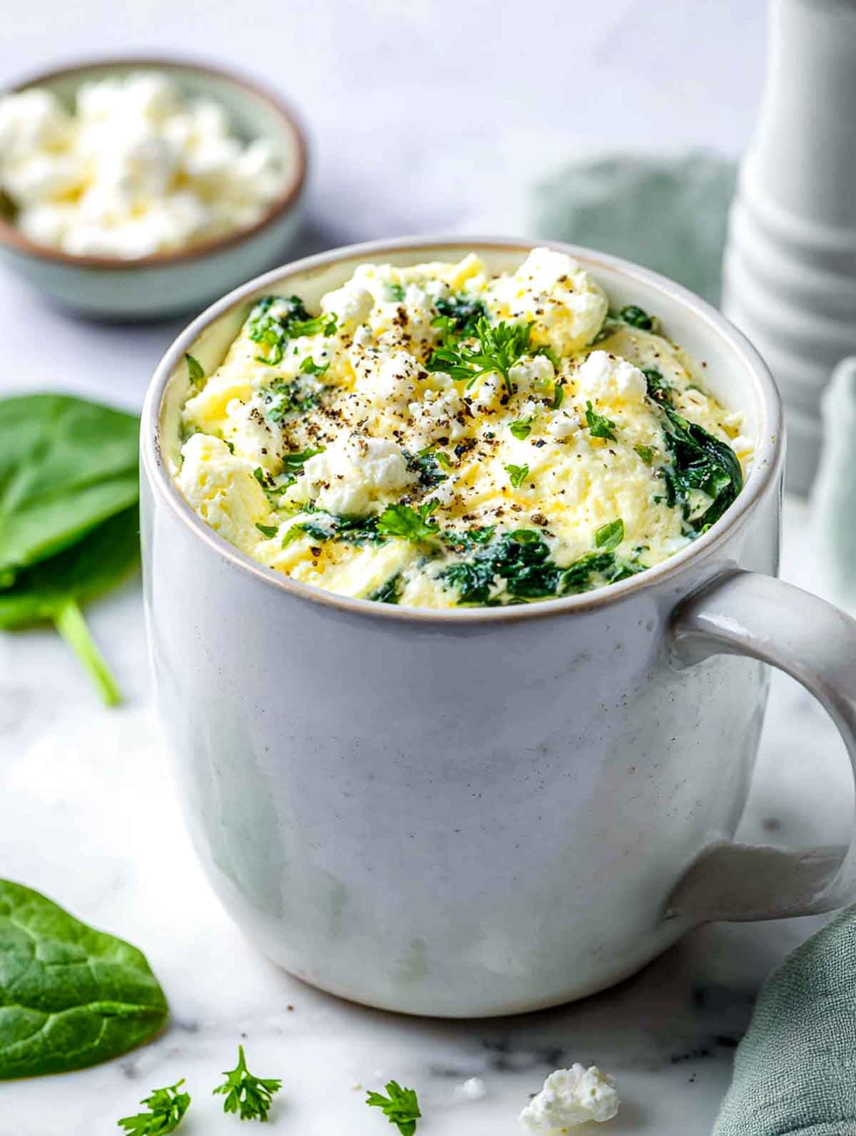 A bright, cool-toned angled side view of a white ceramic mug filled with fluffy spinach feta egg white scramble, topped with crumbled feta, cracked black pepper, and fresh parsley, set on a white marble countertop with a sage green linen napkin and fresh spinach leaves nearby.
