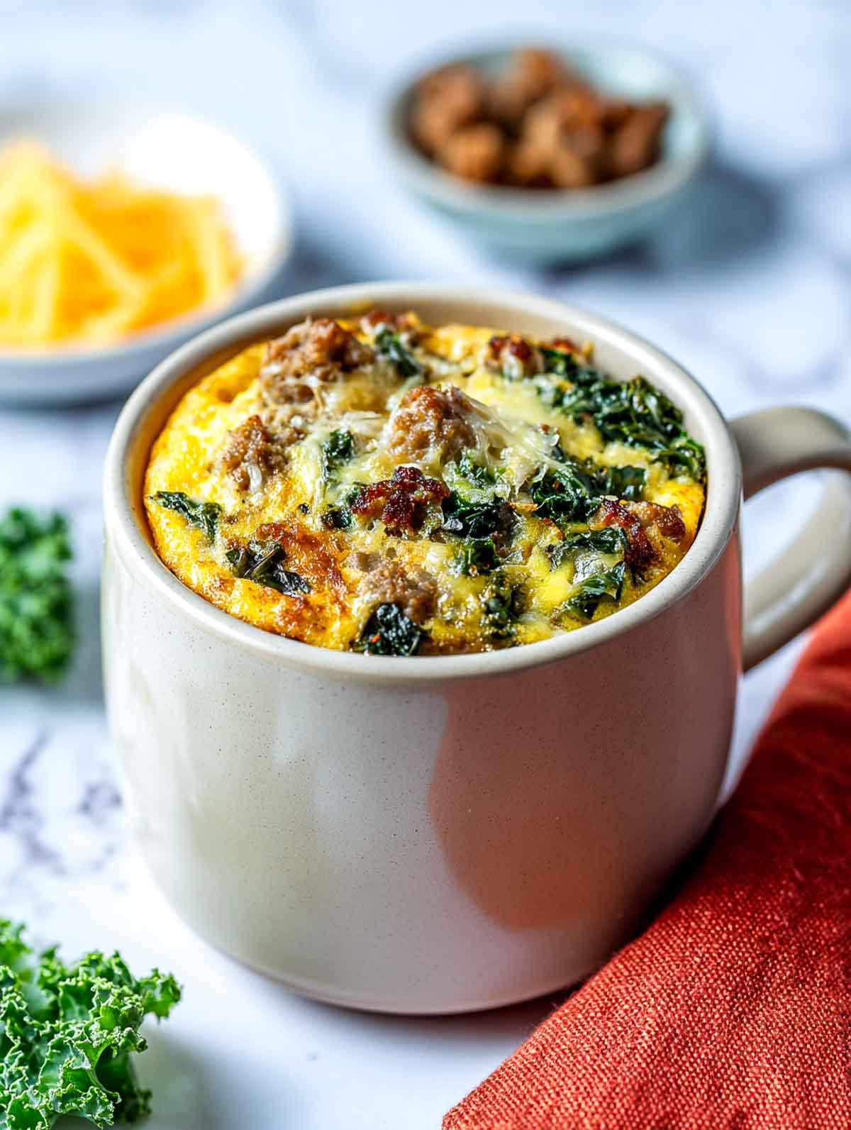 A bright angled view of a cream ceramic mug filled with a turkey sausage and kale egg mug casserole, topped with lightly melted cheese and golden edges, set on a white marble countertop with a rust-colored linen napkin, fresh kale, and small bowls of sausage and shredded cheese nearby.