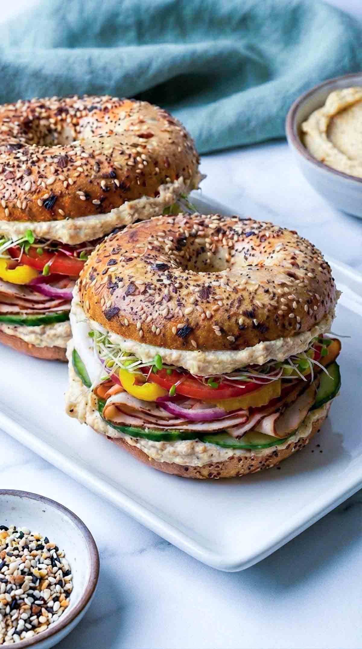 Two everything bagel turkey sandwiches stacked on a white rectangular plate, layered with creamy hummus, folded smoked turkey, cucumber slices, tomato, red onion, banana peppers, and sprouts, with a small bowl of everything seasoning and extra hummus in the background.