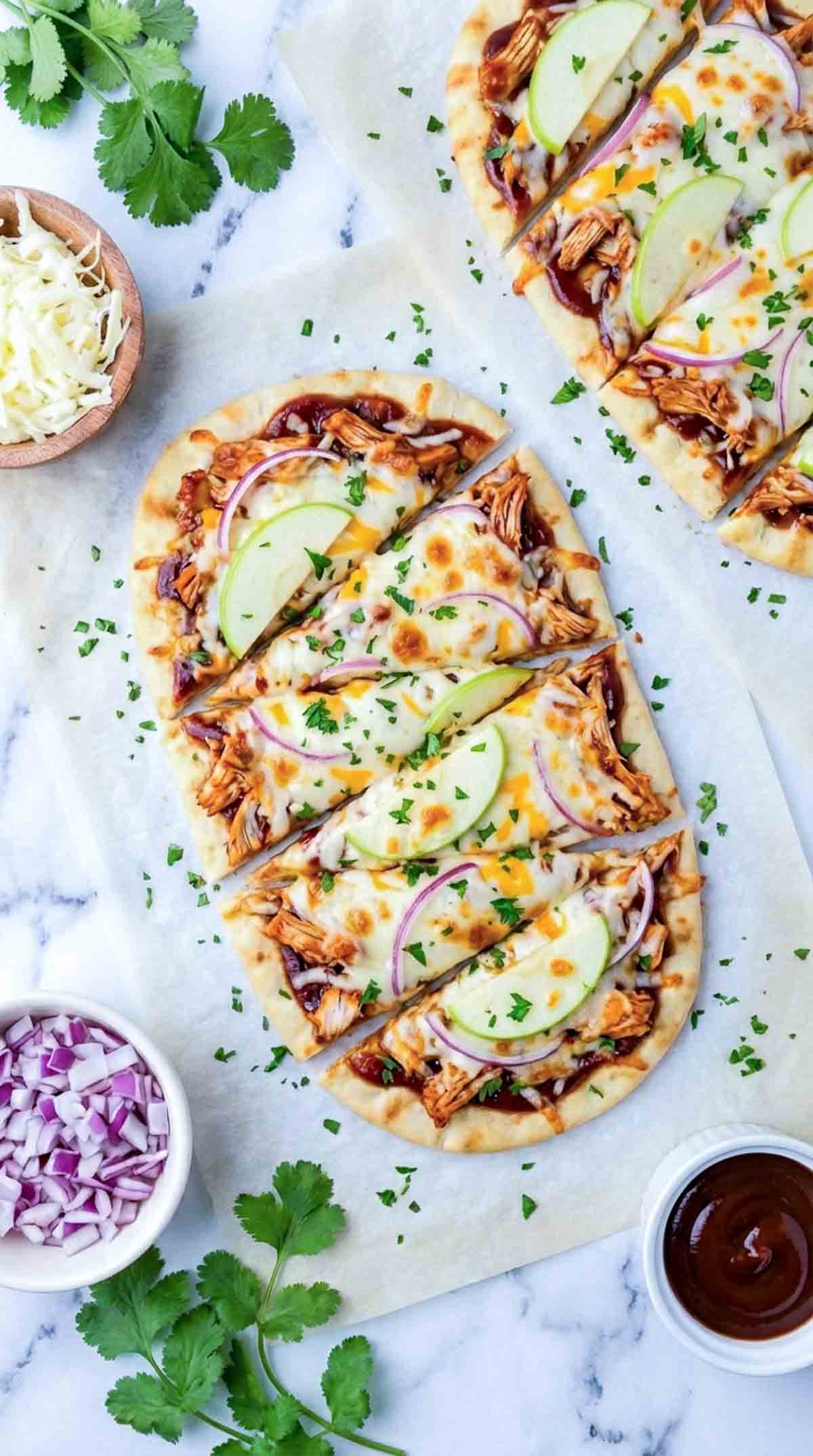 Two sliced BBQ chicken apple cheddar flatbreads on white marble, topped with barbecue sauce, shredded chicken, melted mozzarella and cheddar cheese, thin green apple slices, red onion, and chopped parsley, with bowls of diced red onion and barbecue sauce nearby.