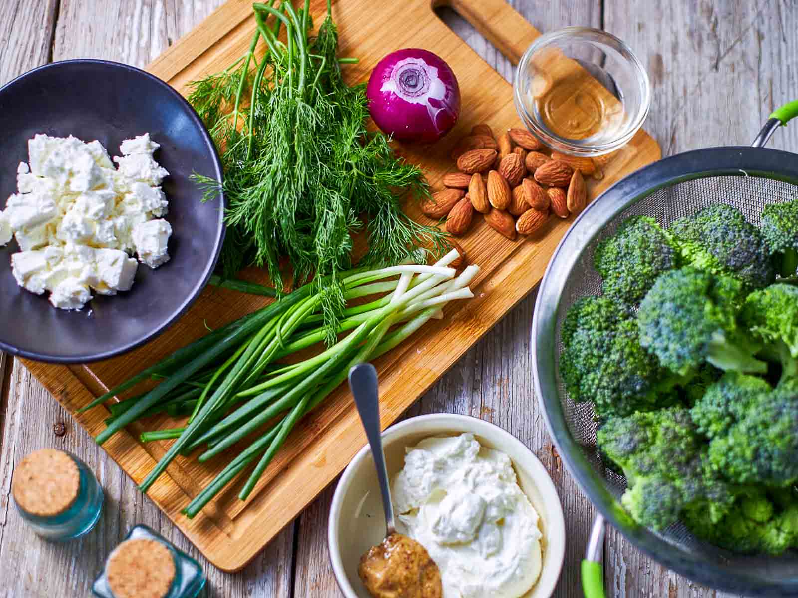 Ingredients for broccoli salad including broccoli, feta cheese, almonds, red onion, herbs, and dressing.