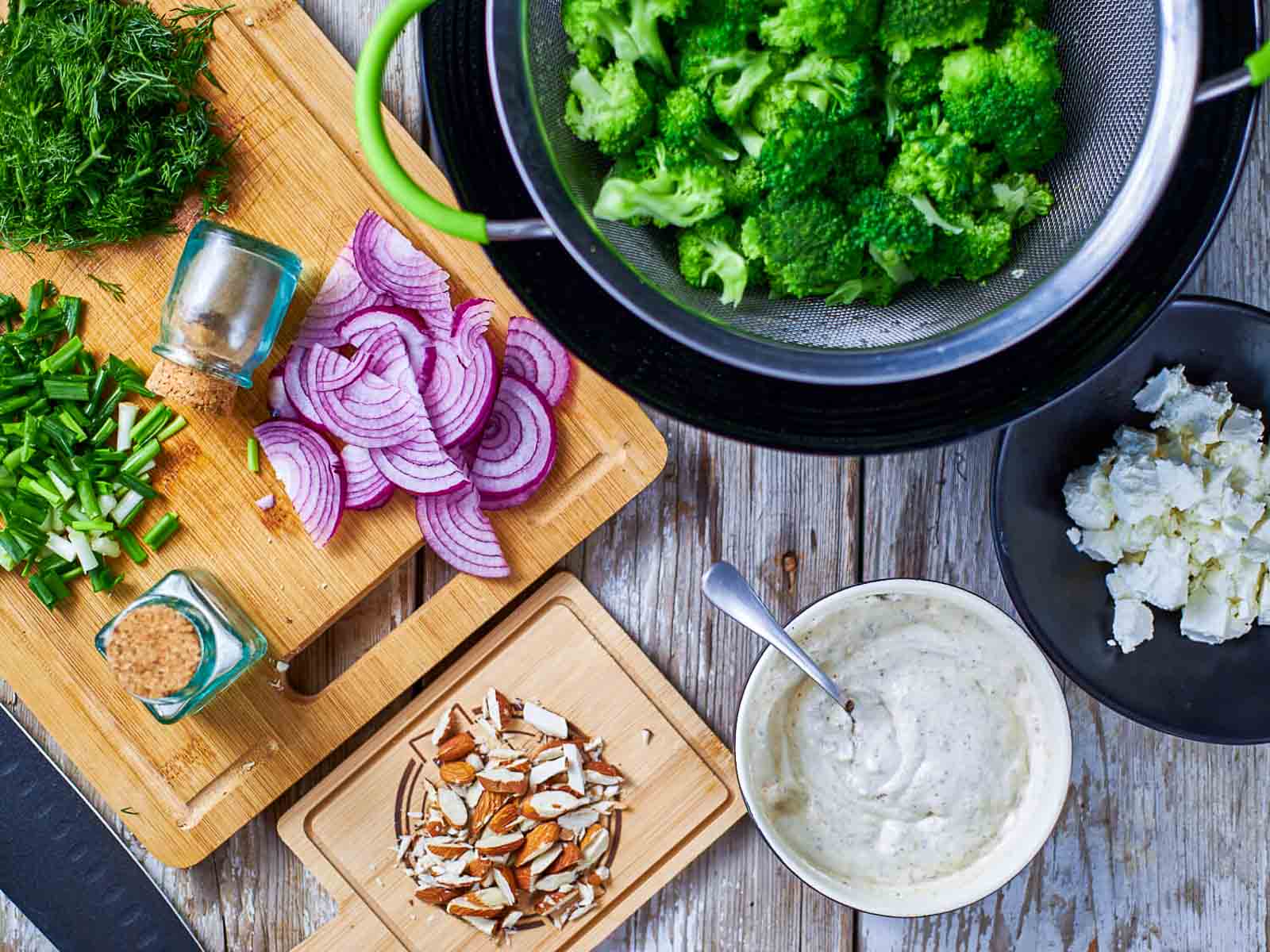 Broccoli florets draining in a colander with sliced red onion and chopped herbs nearby.