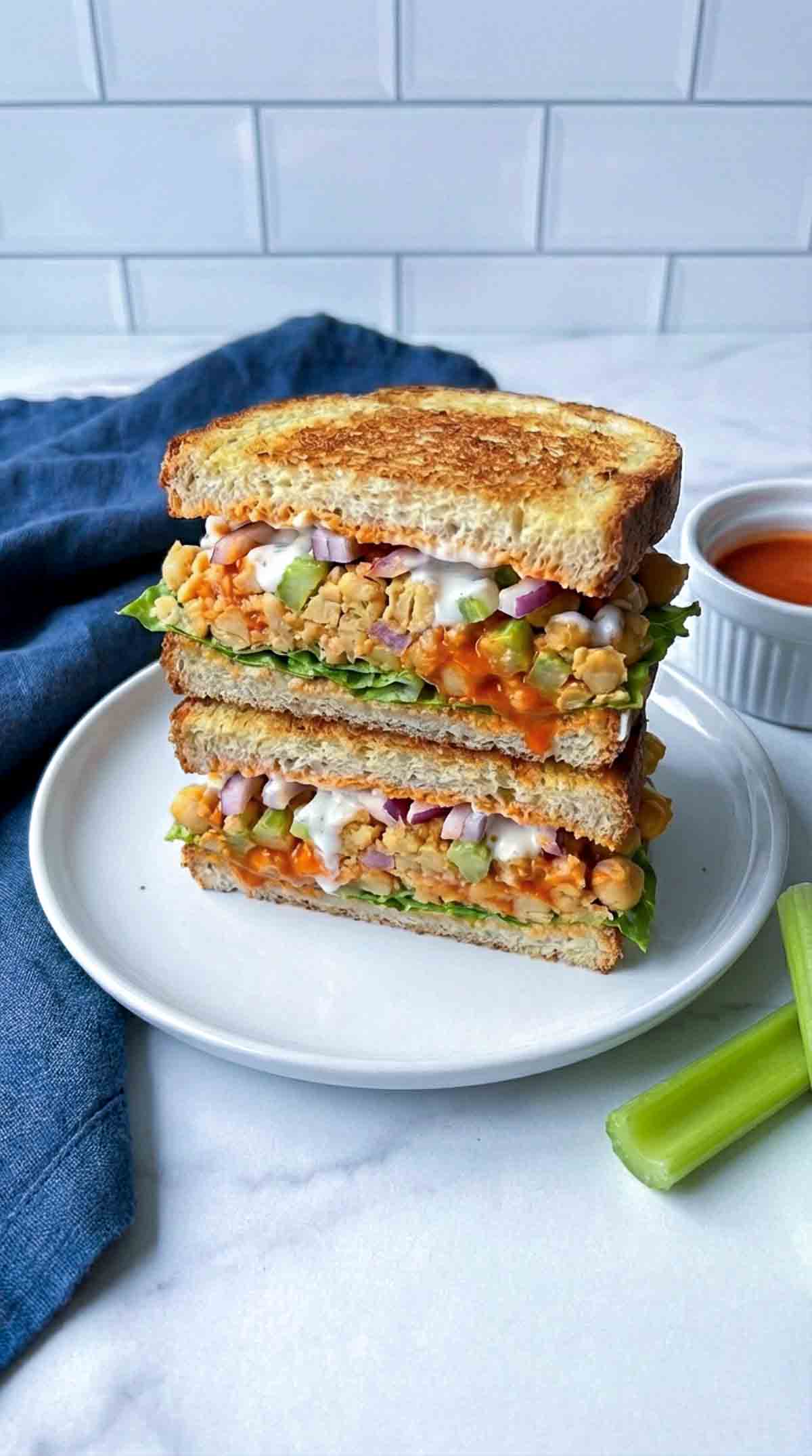 Side view of a buffalo chickpea smash sandwich cut in half and stacked on a white plate, filled with chunky mashed chickpeas coated in orange buffalo sauce, diced celery, red onion, and a drizzle of creamy yogurt sauce, with a small ramekin of buffalo sauce and a celery stick on a white marble surface.
