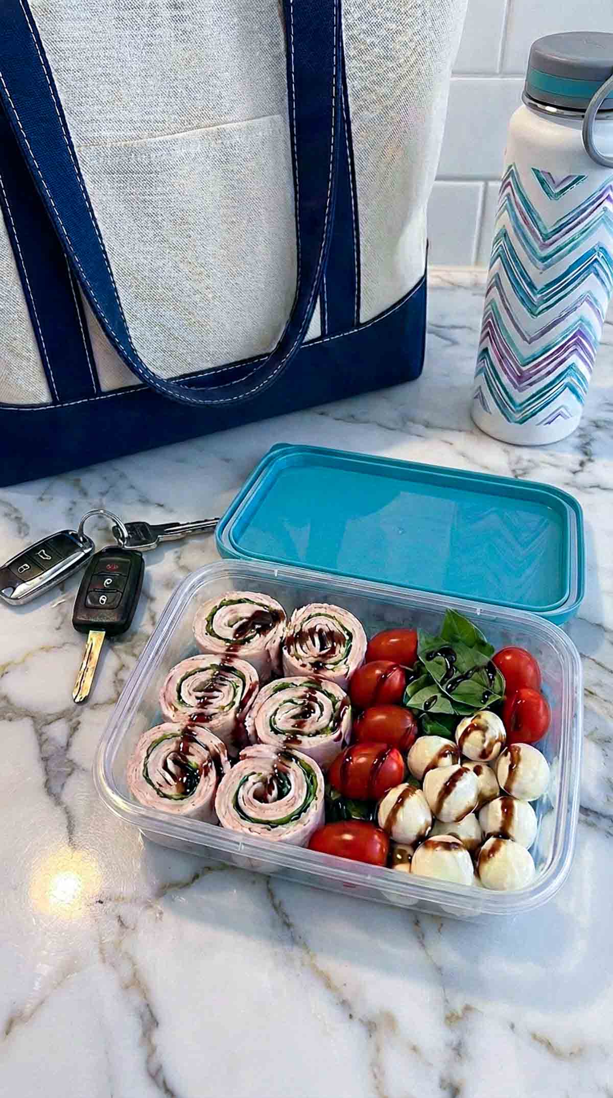 Clear lunch container filled with sliced turkey Caprese wrap pinwheels made with a plain flour tortilla, layered with creamy spread and basil, drizzled with balsamic glaze, alongside cherry tomatoes and mozzarella balls on a white marble surface with a tote bag in the background.