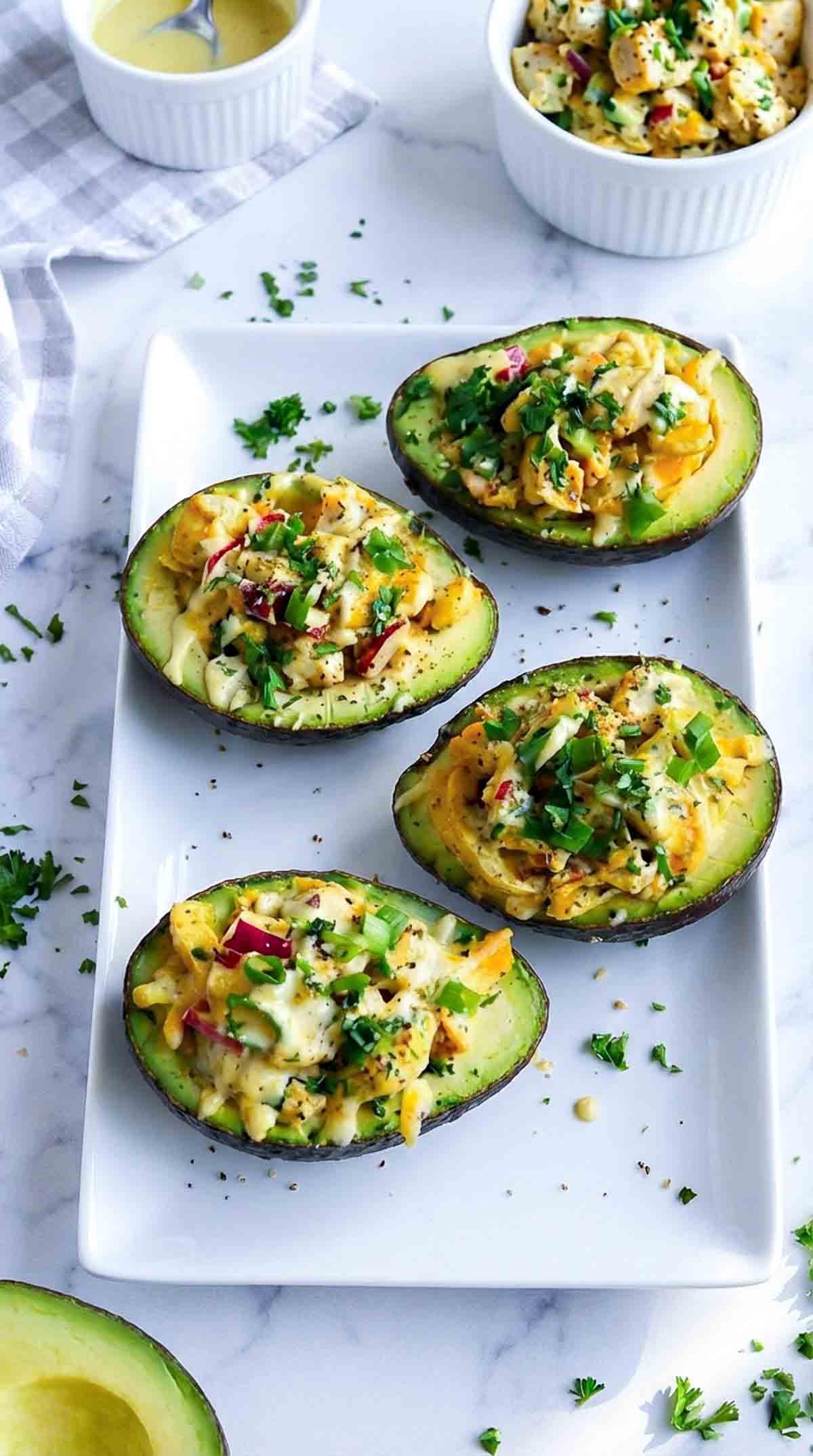 Four avocado halves filled with creamy chicken salad made with shredded chicken, diced celery, red onion, herbs, and dressing, garnished with chopped parsley and cracked black pepper on a white platter over a marble surface.
