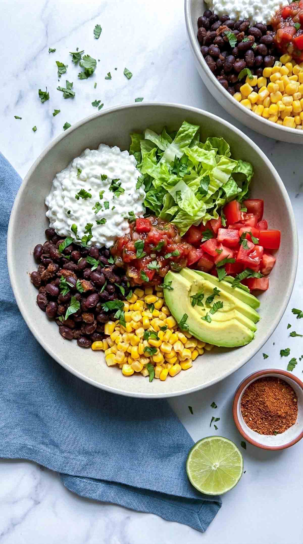 Overhead close-up of a vegetarian cottage cheese taco bowl in a muted beige ceramic bowl on a white marble surface, filled with black beans, sweet corn, diced tomatoes, shredded romaine, sliced avocado, salsa, and a generous scoop of cottage cheese, with chopped cilantro scattered on top, a dusty blue napkin to the side, a lime half, and a small bowl of taco seasoning nearby.