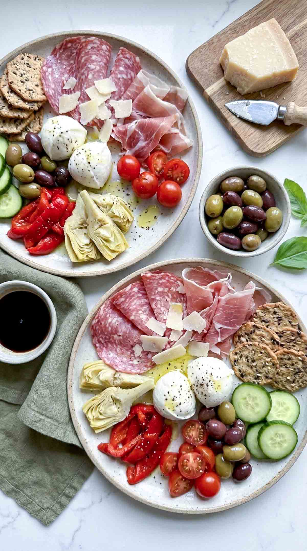 Two Italian antipasto lunch plates with salami, prosciutto, fresh mozzarella, olives, artichokes, roasted red peppers, cherry tomatoes, cucumber slices, crackers, shaved Parmesan, and small bowls of balsamic and olives on a white marble surface.