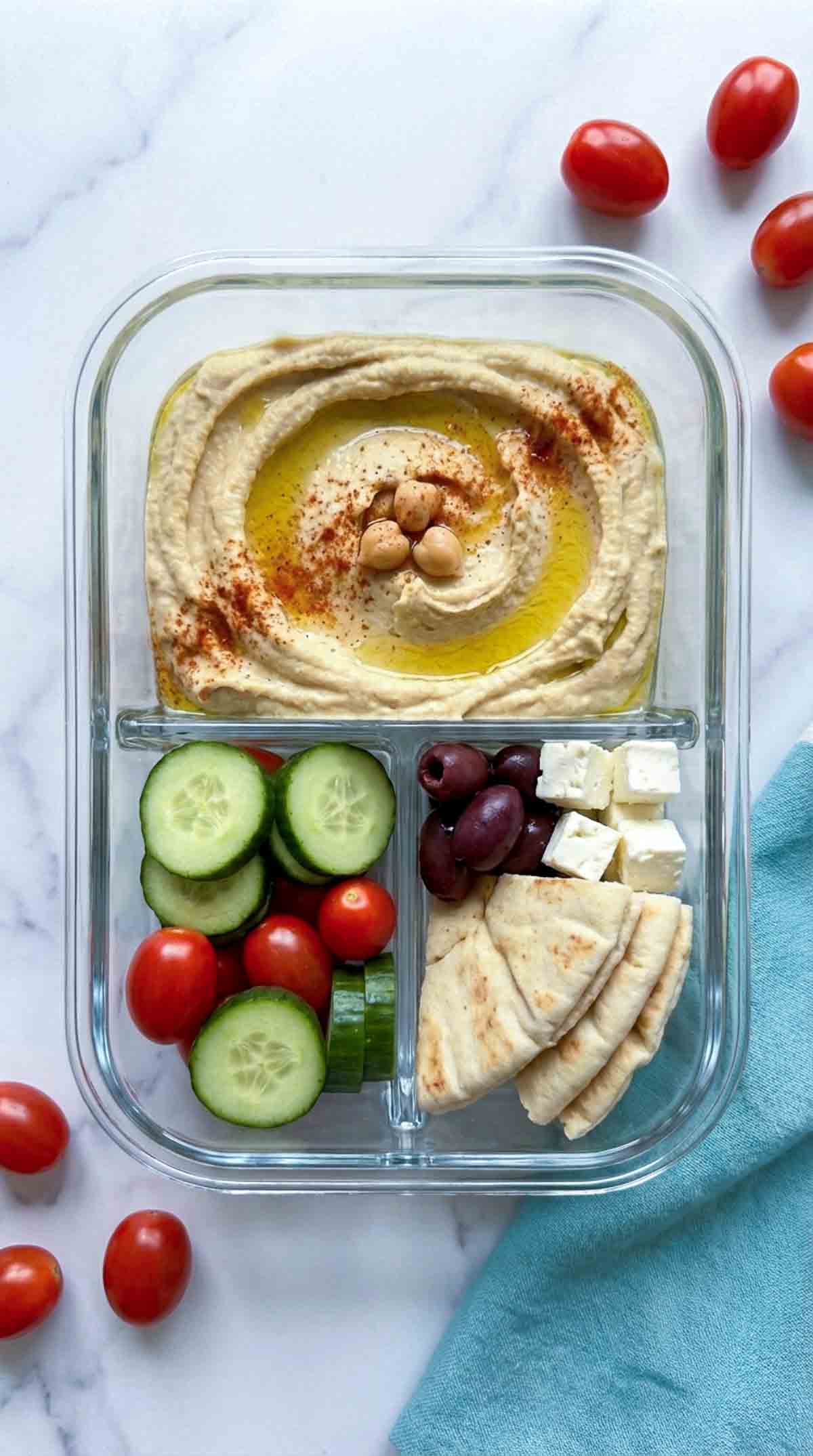 Three clear glass divided meal prep containers arranged diagonally on a white marble surface, each filled with creamy hummus topped with olive oil and paprika, sliced cucumbers, cherry tomatoes, kalamata olives, feta cubes, and folded pita wedges, with a teal linen napkin tucked along the bottom edge and a small bowl of olives at the top.
