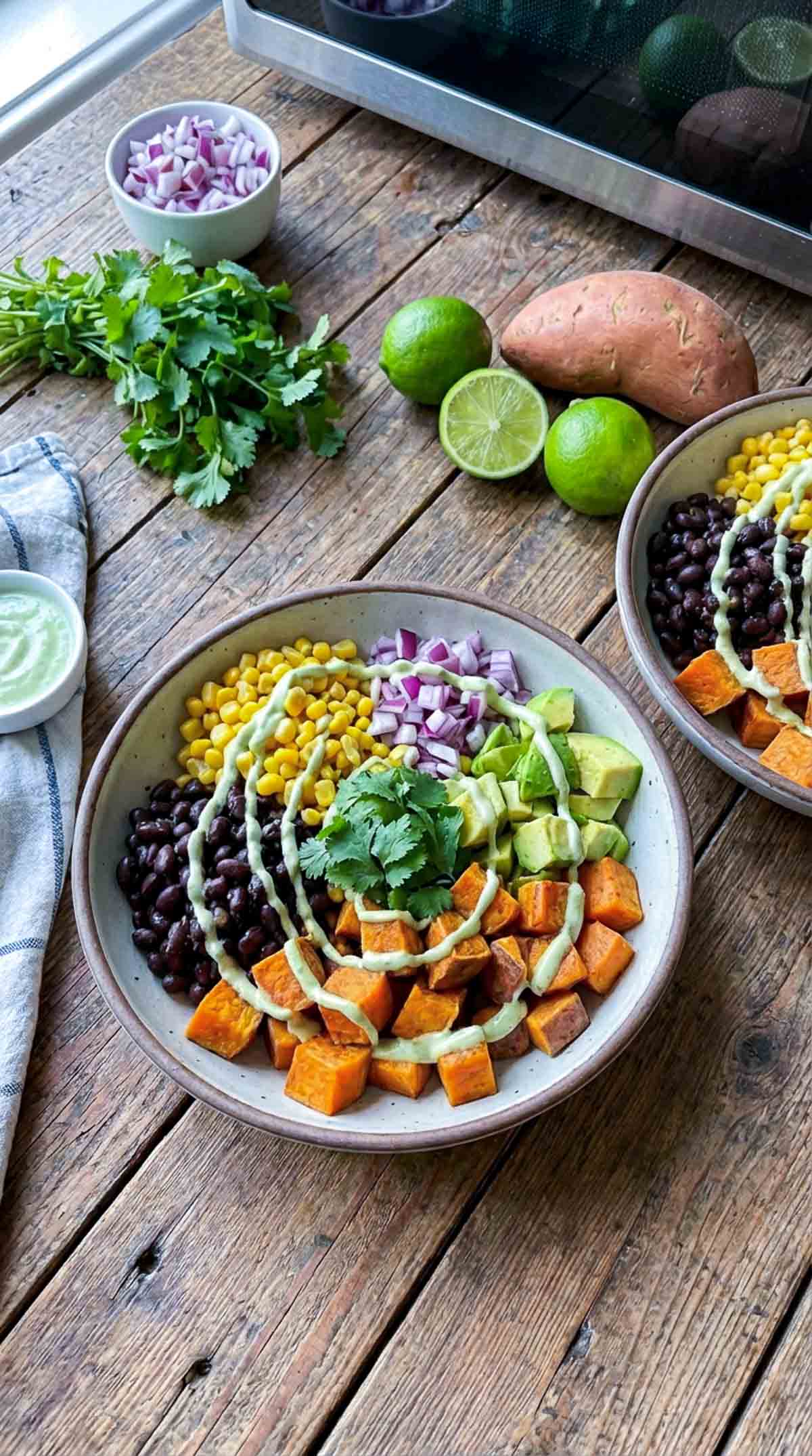 Ceramic bowl filled with microwave sweet potato cubes, black beans, corn, diced red onion, avocado, and cilantro topped with creamy green sauce on a rustic wooden table with limes and fresh cilantro nearby.