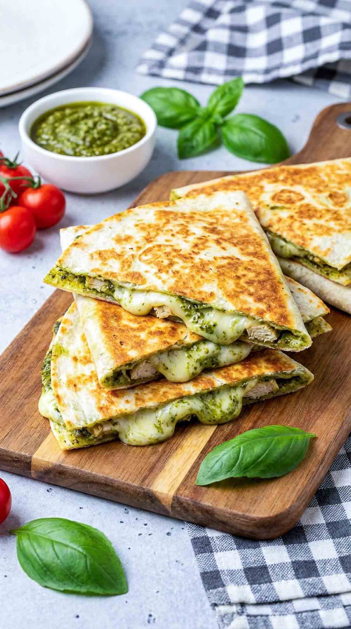 Golden brown chicken pesto quesadilla triangles stacked on a wooden board with melted mozzarella, bright green pesto, fresh basil leaves, cherry tomatoes, and a small bowl of pesto on the side.