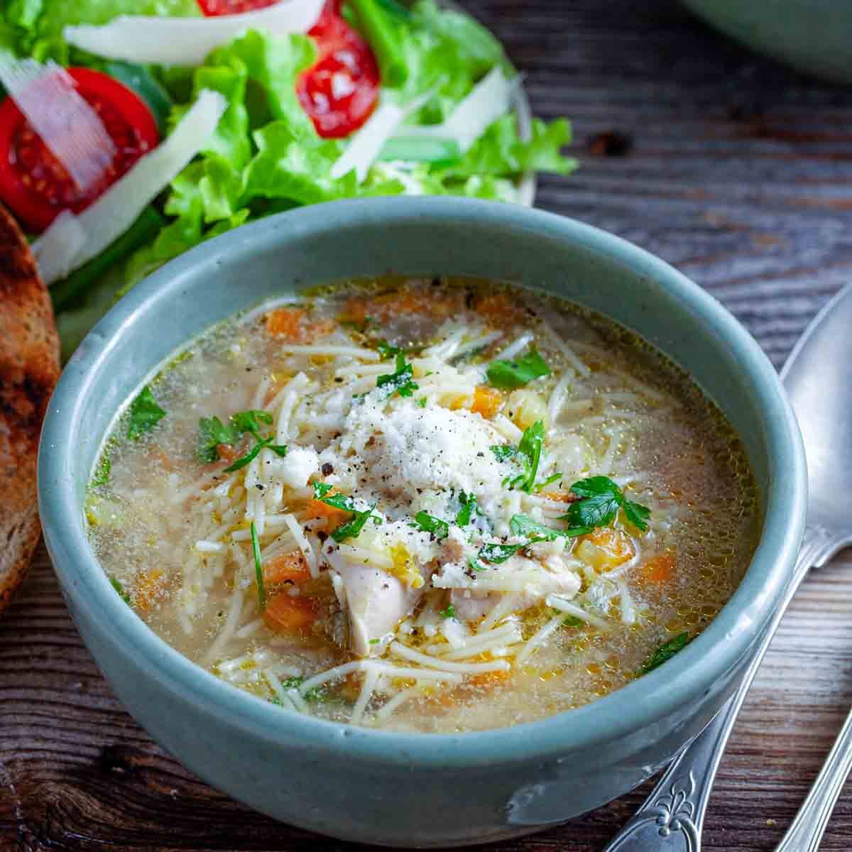 Bowl of chicken fideo soup topped with shredded cheese and parsley, served with bread and salad.