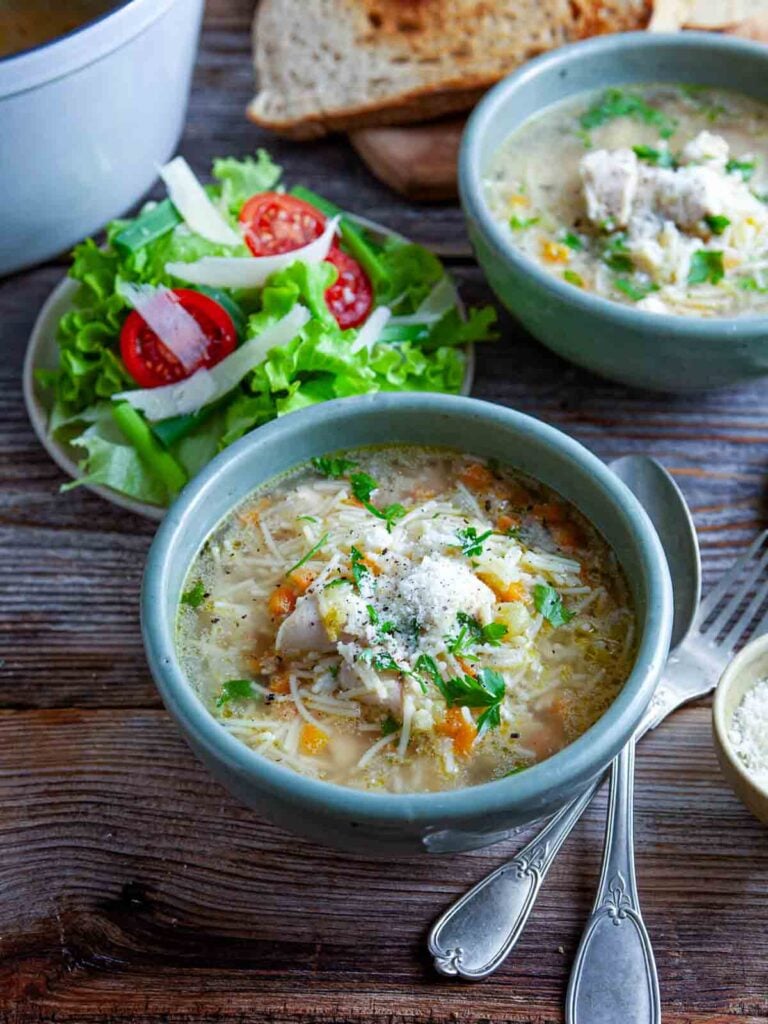 A bowl of San Marino chicken fideo soup topped with shredded chicken, parmesan, and fresh herbs, served with toast and a side salad.