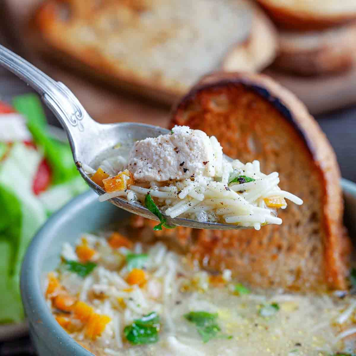 Close-up spoonful of San Marino chicken fideo soup with tender chicken, fideo noodles, carrots, and herbs, with toasted bread in the background.