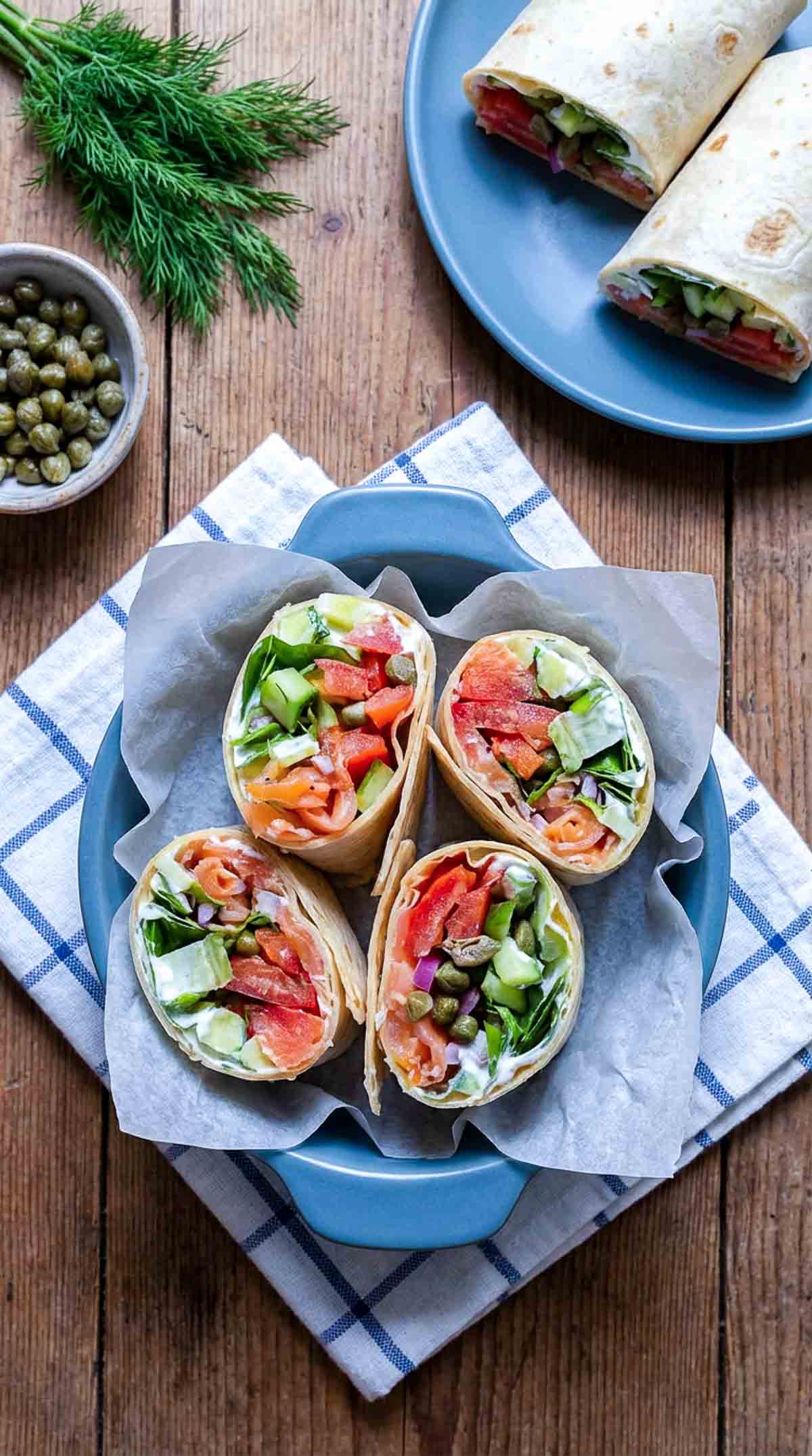 Four smoked salmon wrap halves arranged upright in a blue ceramic dish lined with parchment paper, showing layers of smoked salmon, diced cucumber, tomato, red onion, romaine lettuce, capers, and cream cheese inside soft flour tortillas on a wooden surface.