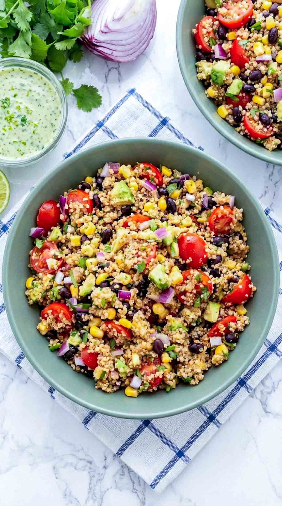 Two wide green ceramic bowls filled with fluffy quinoa mixed with black beans, corn, halved cherry tomatoes, diced avocado, red onion, and cilantro, served on a white marble surface with a white and blue check napkin and a small bowl of creamy cilantro lime dressing on the side.