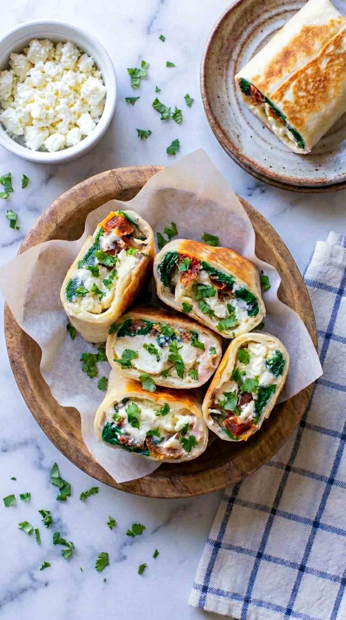 Four toasted spinach feta egg wrap halves stacked in a wooden bowl lined with parchment paper, showing fluffy egg, wilted spinach, sun-dried tomatoes, and melted feta inside lightly browned flour tortillas on a white marble surface with a small bowl of feta and a linen napkin nearby.