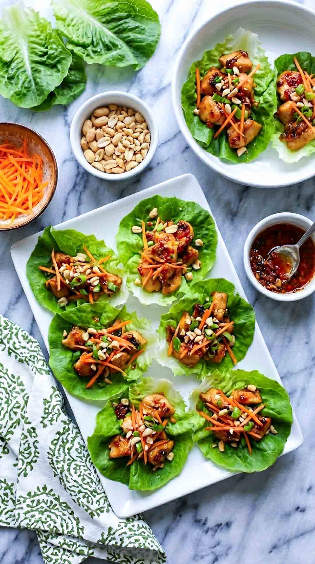 Six teriyaki chicken lettuce cups arranged on a white square plate over white marble, filled with glossy diced chicken, shredded carrots, chopped green onions, and crushed peanuts, with bowls of carrots, peanuts, extra teriyaki sauce, and fresh lettuce leaves nearby.