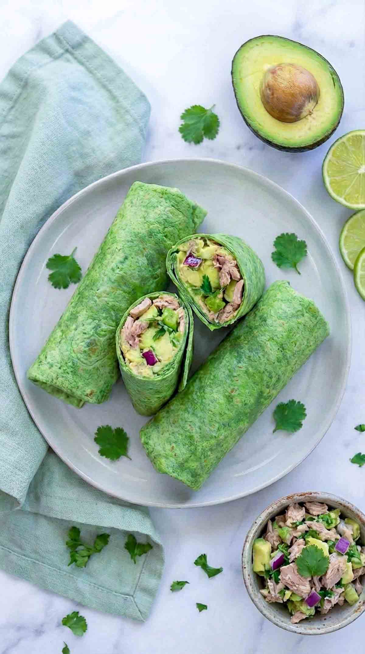 Overhead view of four spinach tortilla avocado tuna wraps arranged on a muted light ceramic plate on a white marble surface, two whole wraps and two sliced in half showing chunky tuna, diced avocado, red onion, and herbs, with a light green linen napkin, a halved avocado, and a lime wedge beside the plate.
