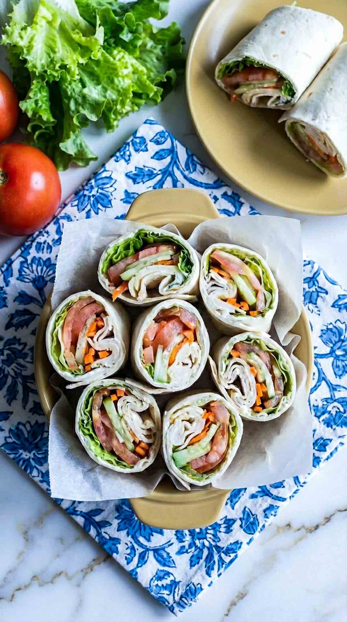 Four fresh veggie cream cheese wraps sliced in half and arranged upright in a shallow dish lined with parchment paper, filled with herbed cream cheese, crisp lettuce, tomato slices, shredded carrots, thin red bell pepper strips, and provolone cheese, set on white marble with a blue patterned napkin.
