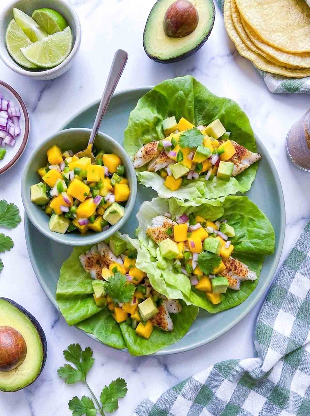 Fish taco lettuce wraps placed on a green serving plate with a bowl of mango salsa next to them.