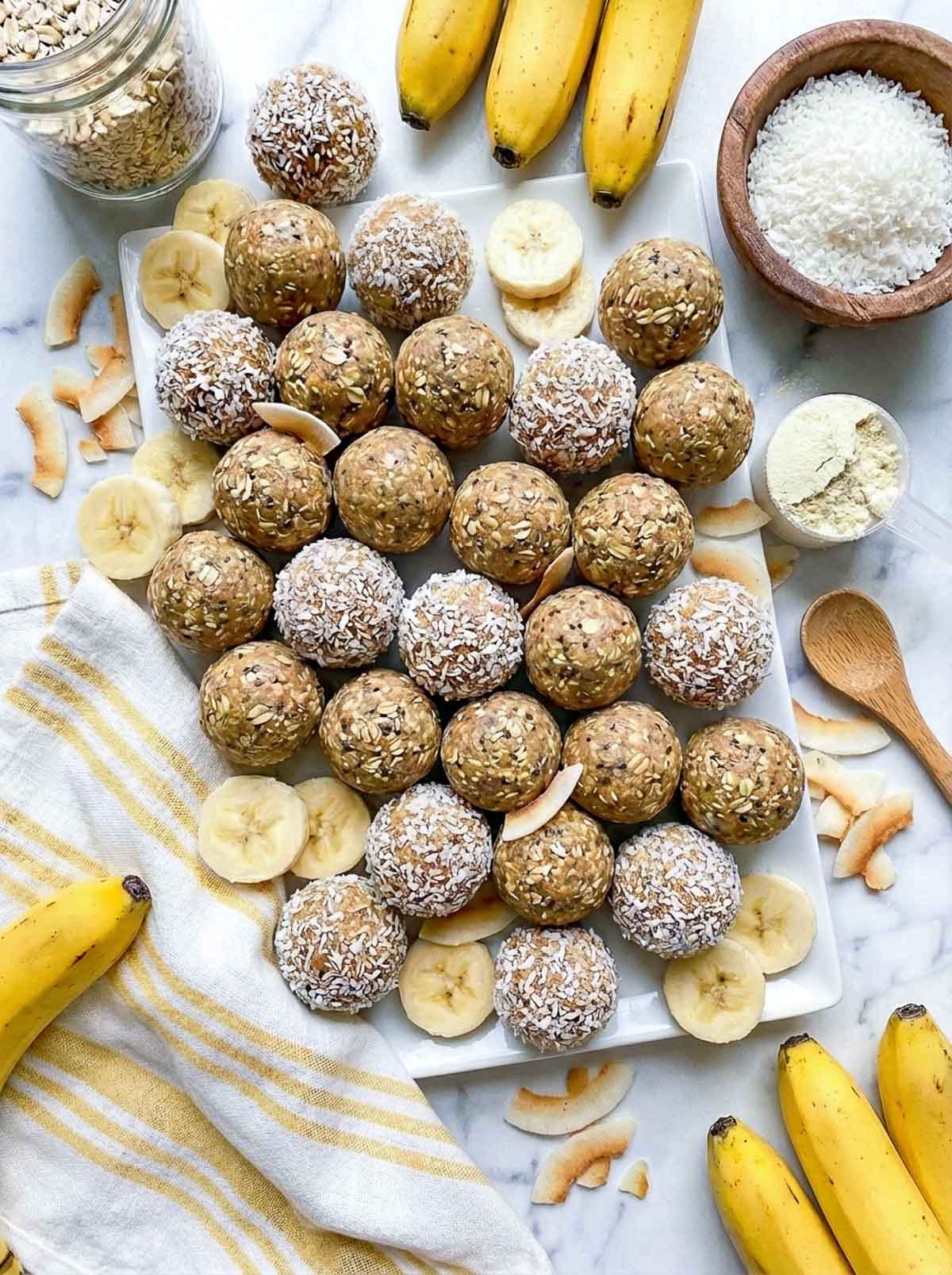 Healthy banana coconut high-protein energy balls on a white rectangular tray, some rolled in shredded coconut and some left plain, surrounded by fresh banana slices, whole bananas, toasted coconut flakes, a wooden bowl of shredded coconut, and a scoop of protein powder on a marble surface.