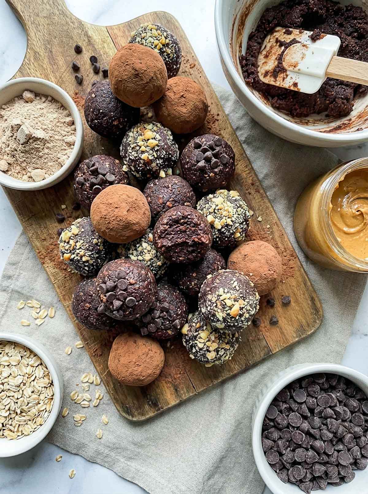 No-bake brownie batter high-protein energy balls on a wooden board finished three ways — dusted with cocoa powder, rolled in crushed nuts, and topped with chocolate chips — with a mixing bowl of brownie batter, protein powder, peanut butter, oats, and chocolate chips surrounding them.