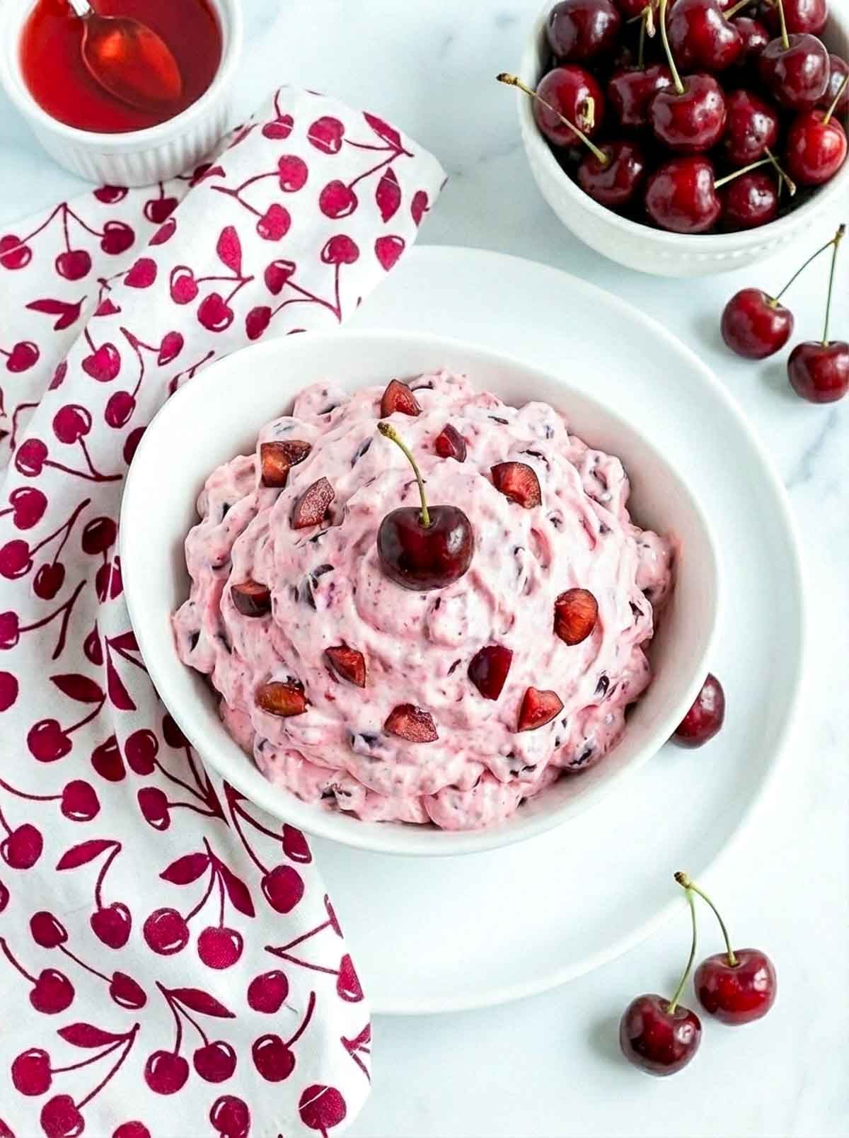 Cherry cheesecake fluff in a white bowl with a creamy, fluffy texture and cherry pieces, surrounded by fresh cherries and a patterned napkin.