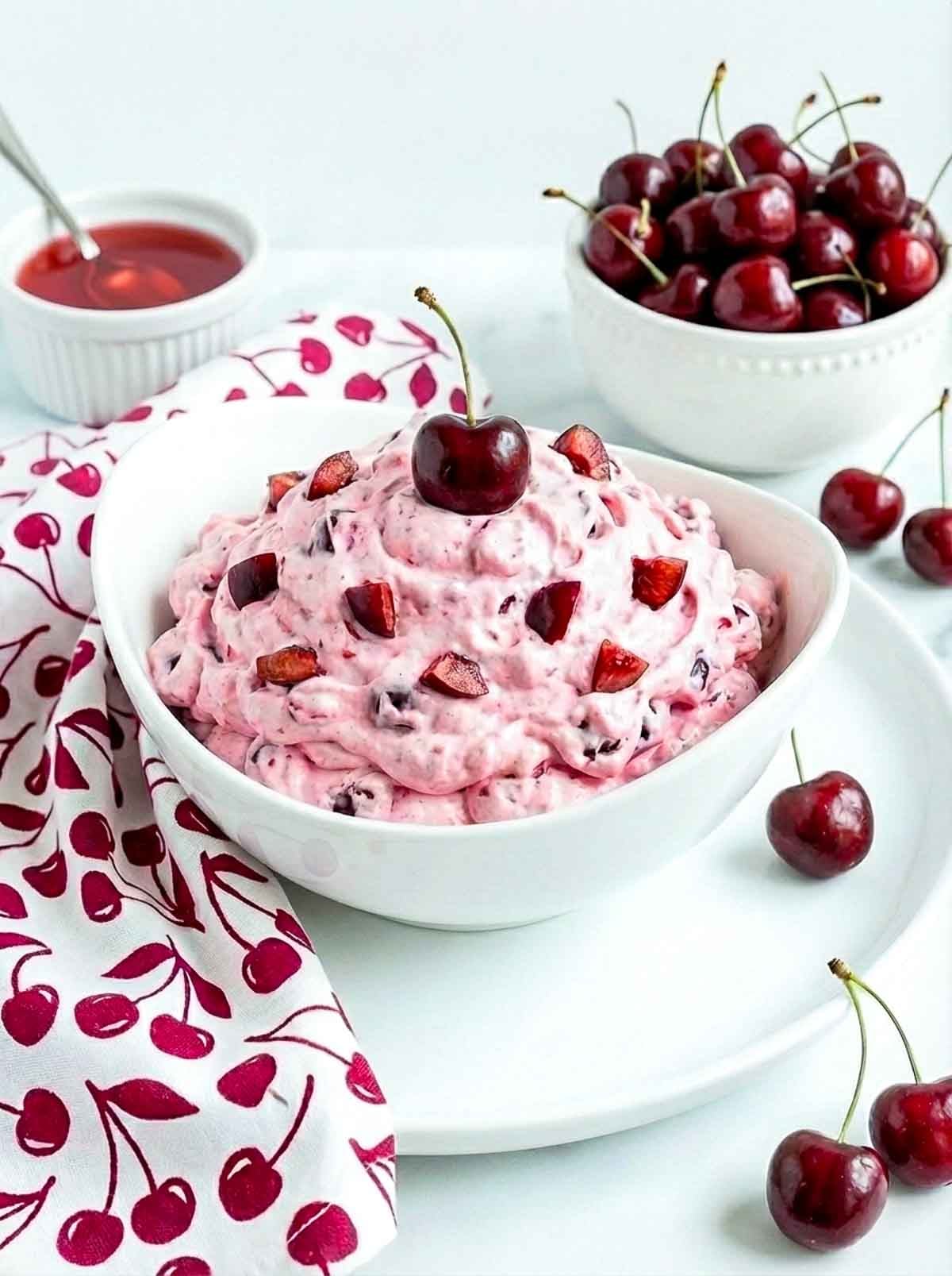 A bowl of cherry cheesecake fluff topped with diced cherries and a whole cherry, set on a white plate with fresh cherries and a cherry-patterned napkin.