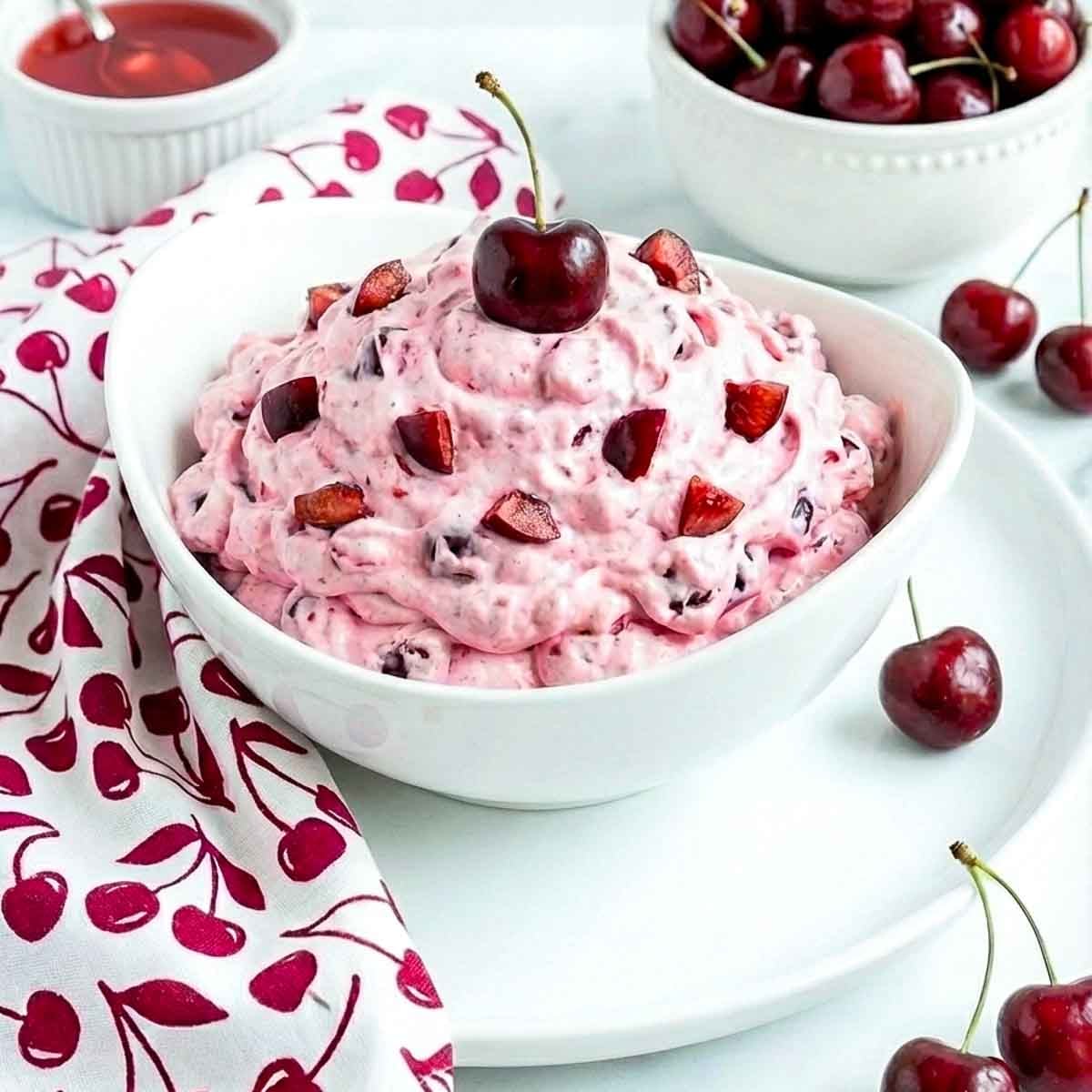 Close-up of cherry cheesecake fluff in a bowl with diced cherries and a whole cherry on top, styled with fresh cherries and a cherry-print napkin.