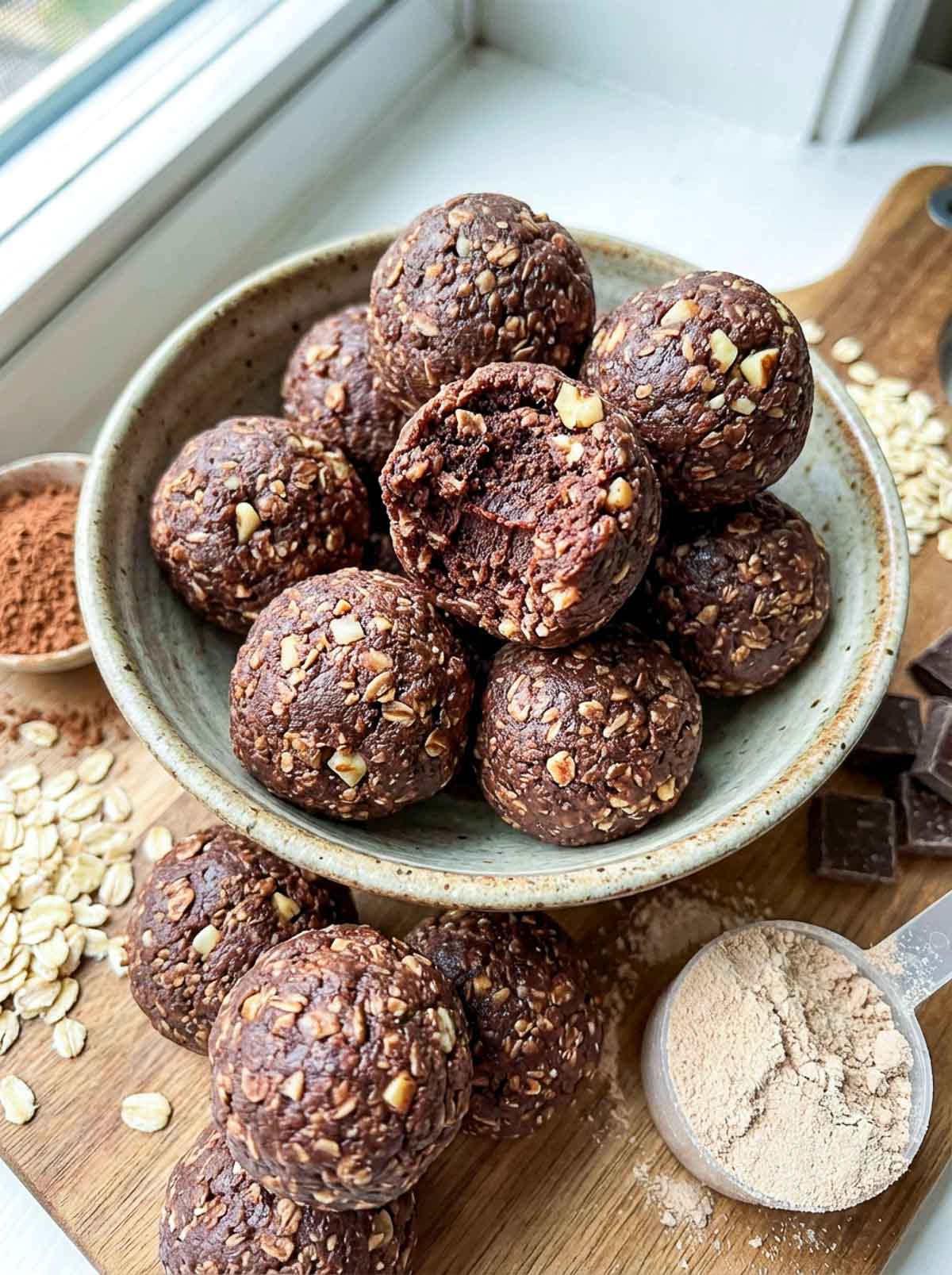 Chocolate brownie oat high-protein energy balls in a speckled ceramic bowl on a wooden board, with one ball bitten open showing a fudgy interior, surrounded by rolled oats, chunks of dark chocolate, a bowl of cocoa powder, and a scoop of protein powder.