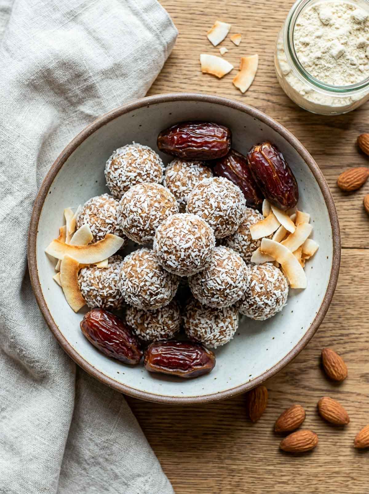 Vegan date-sweetened coconut high-protein energy balls rolled in shredded coconut in a speckled ceramic bowl, surrounded by whole medjool dates, raw almonds, and toasted coconut flakes on a wooden surface.