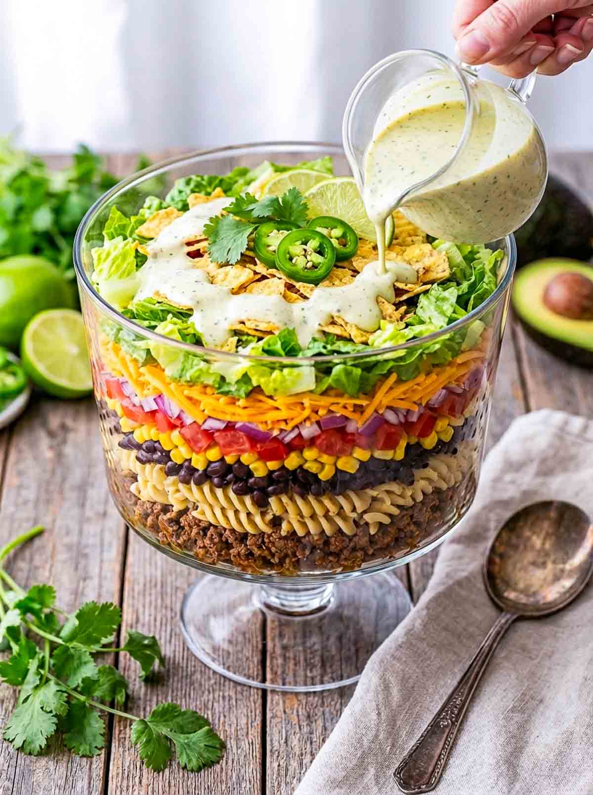 Side view of a seven layered pasta salad with lime cilantro dressing being poured on it.