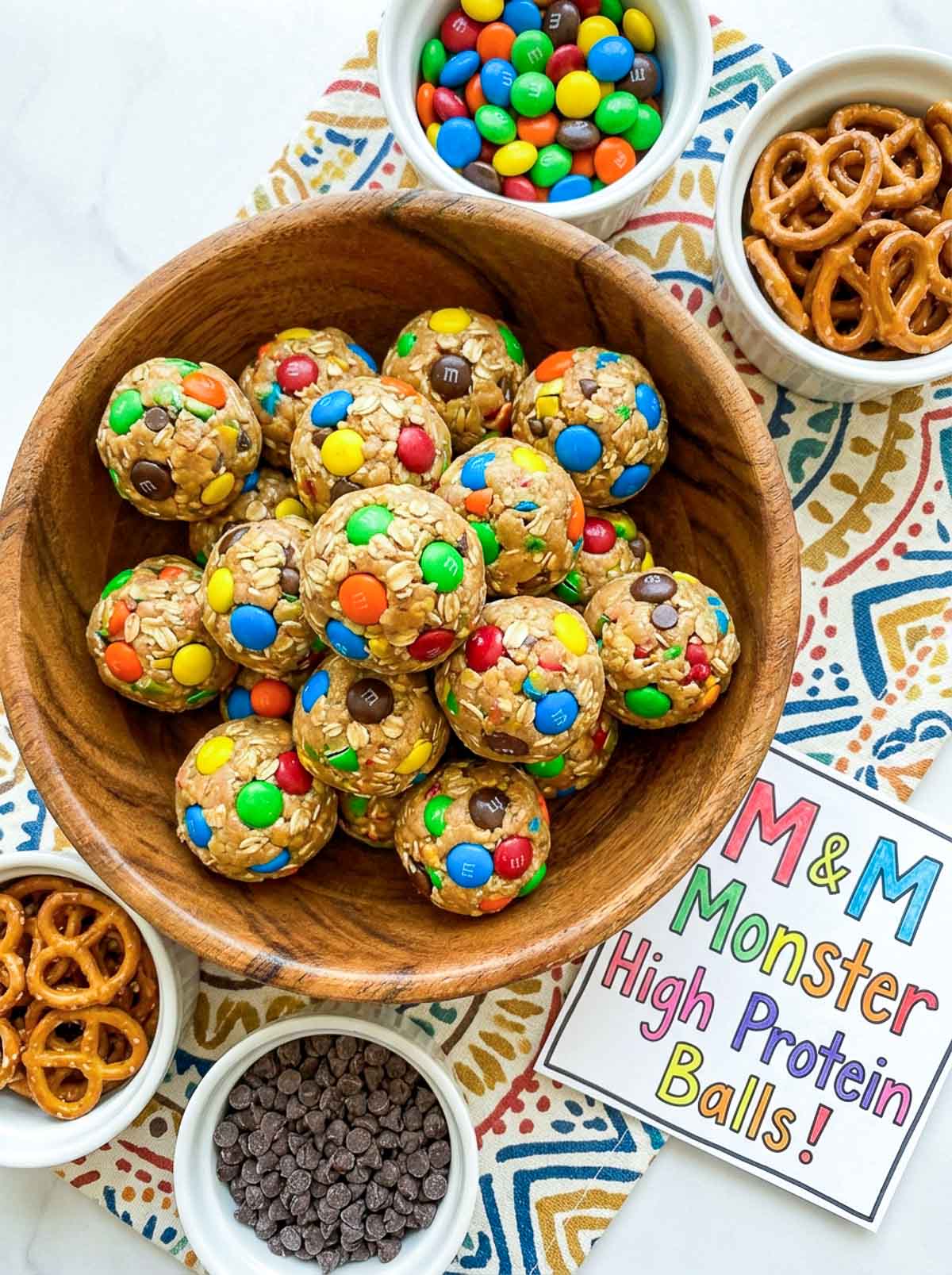 M&M monster high-protein energy balls for kids packed with colorful M&Ms in a wooden bowl, surrounded by bowls of M&Ms, mini pretzels, and chocolate chips on a colorful patterned cloth.