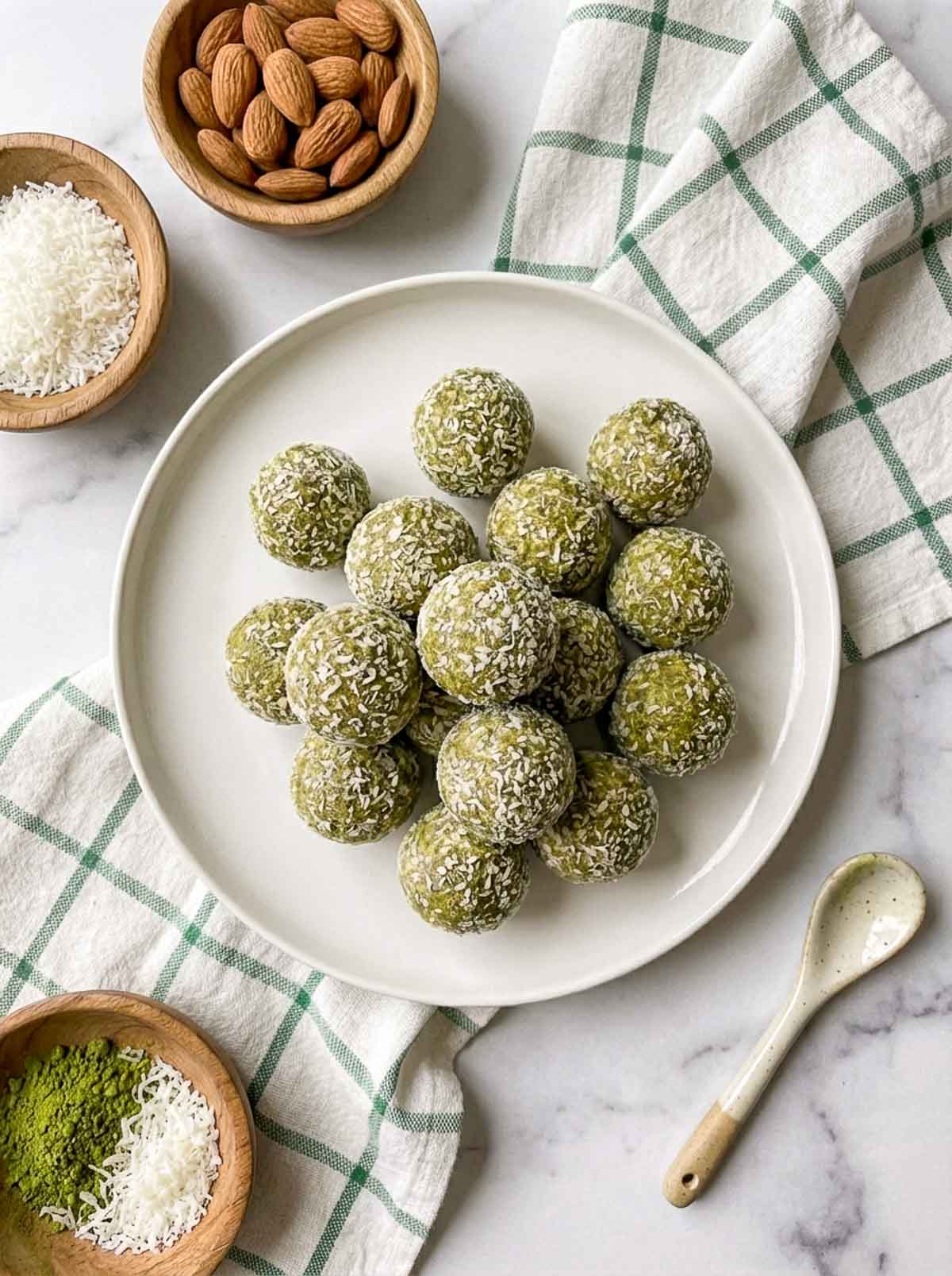 Matcha coconut high-protein energy balls rolled in shredded coconut on a white plate, surrounded by wooden bowls of matcha powder, shredded coconut, and raw almonds on a marble surface with a green checkered cloth.