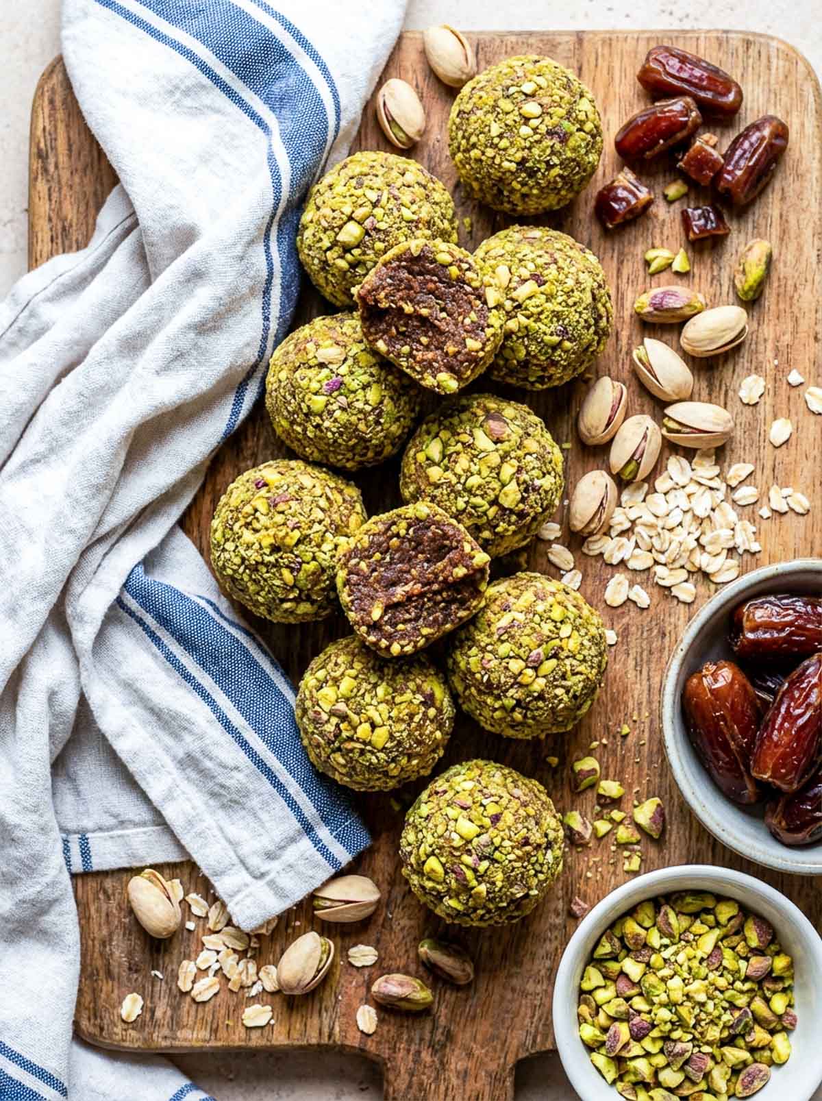 Healthy pistachio date high-protein energy balls rolled in finely chopped pistachios on a rustic wooden board, with two balls cut open to show the dark date interior, whole medjool dates, pistachios, rolled oats, and a small bowl of chopped pistachios alongside a blue striped cloth.