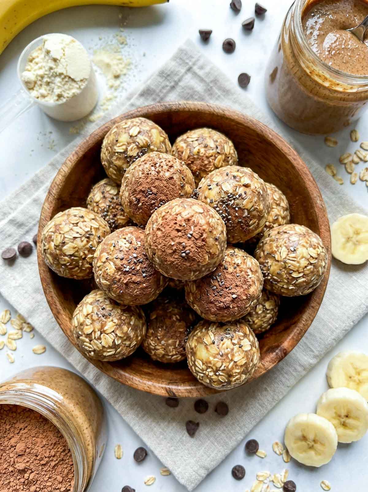 Protein powder banana oat high-protein energy balls in a wooden bowl, some dusted with cocoa powder and chia seeds on top, surrounded by sliced banana, scattered oats, chocolate chips, a scoop of protein powder, and jars of almond butter on a light surface.
