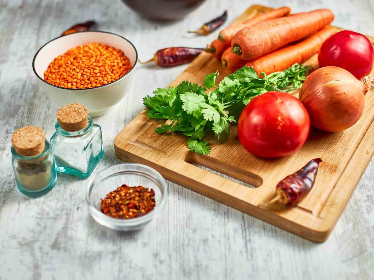 Fresh ingredients like carrots, tomatoes, onion, cilantro, red lentils, and spices are arranged on a board, likely for making a flavorful spiced soup.