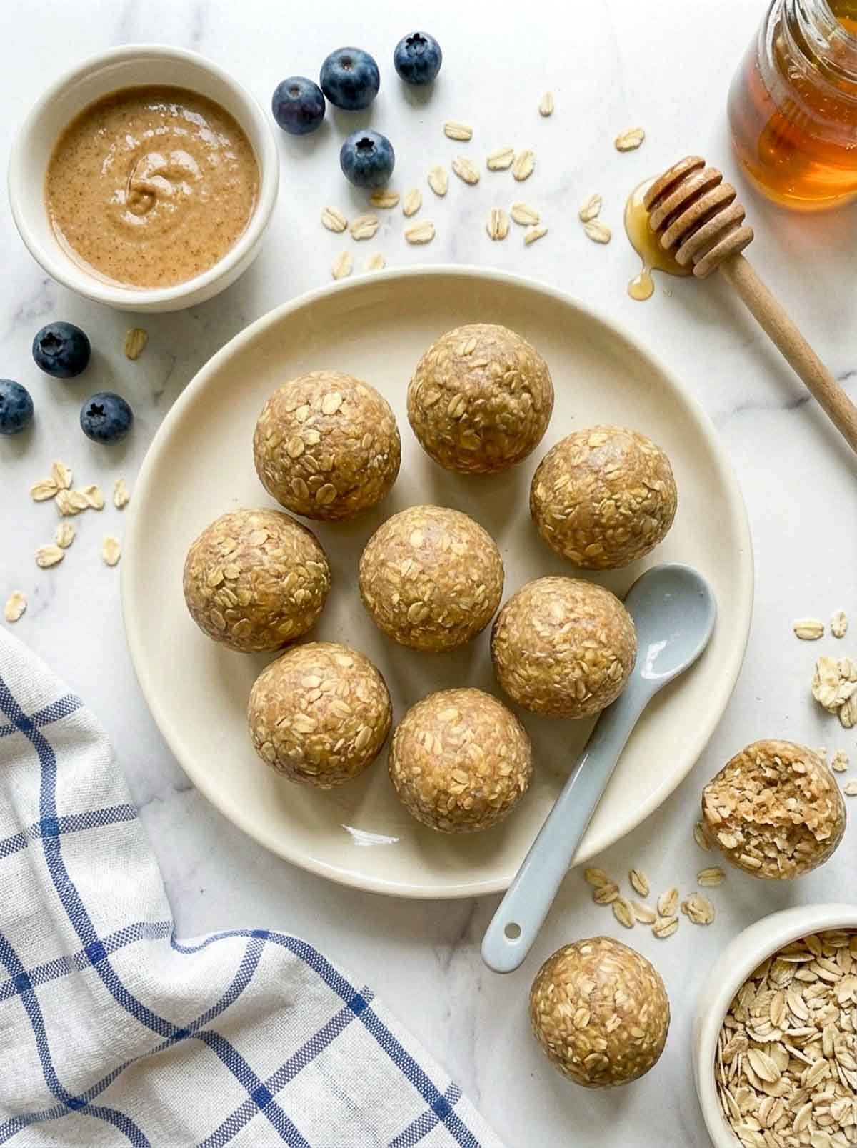 Toddler-friendly oatmeal high-protein energy balls on a cream ceramic plate with a small spoon, surrounded by fresh blueberries, a bowl of almond butter, a honey dipper, a jar of honey, and a bowl of rolled oats on a marble surface with a blue checkered cloth.