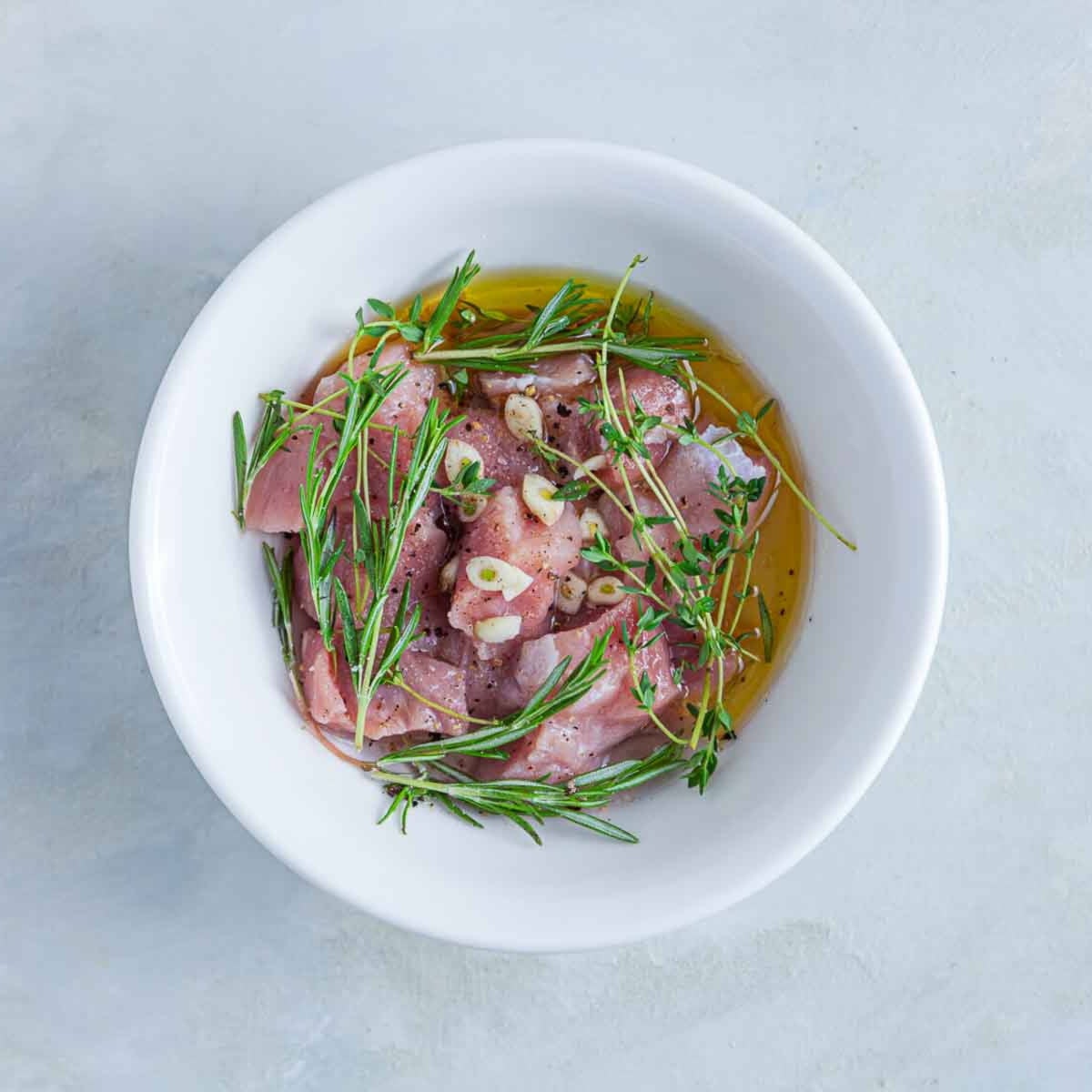 Raw meat marinating with olive oil, garlic slices, and fresh herbs like rosemary and thyme