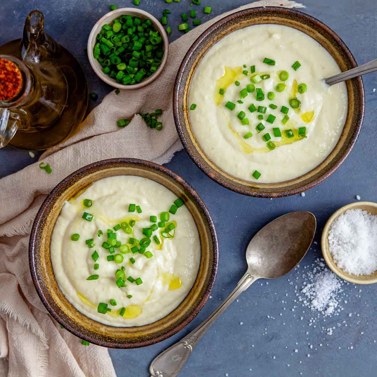 Two bowls of creamy soup topped with chopped green onions and a drizzle of olive oil, served with salt and utensils nearby